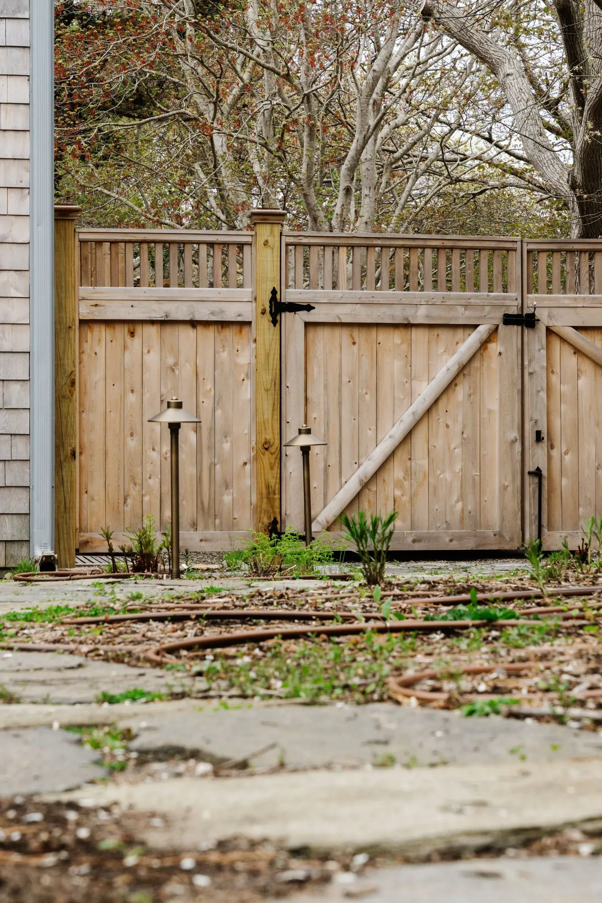 Wooden backyard gate and fence beside a stone patio, with trees and fallen leaves in the background