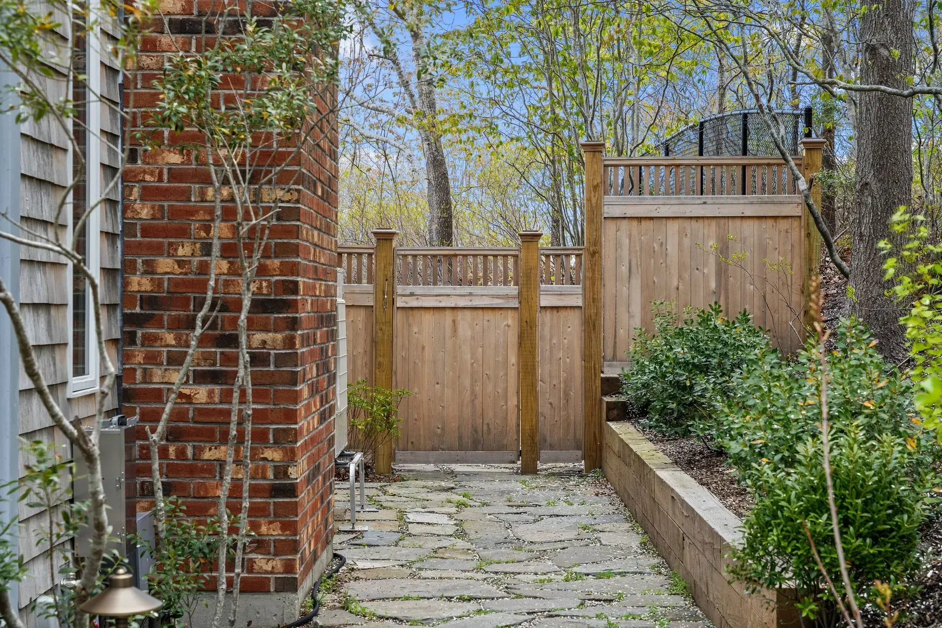 Narrow stone path beside a brick wall and wooden fence, with trees and shrubs in a backyard.
