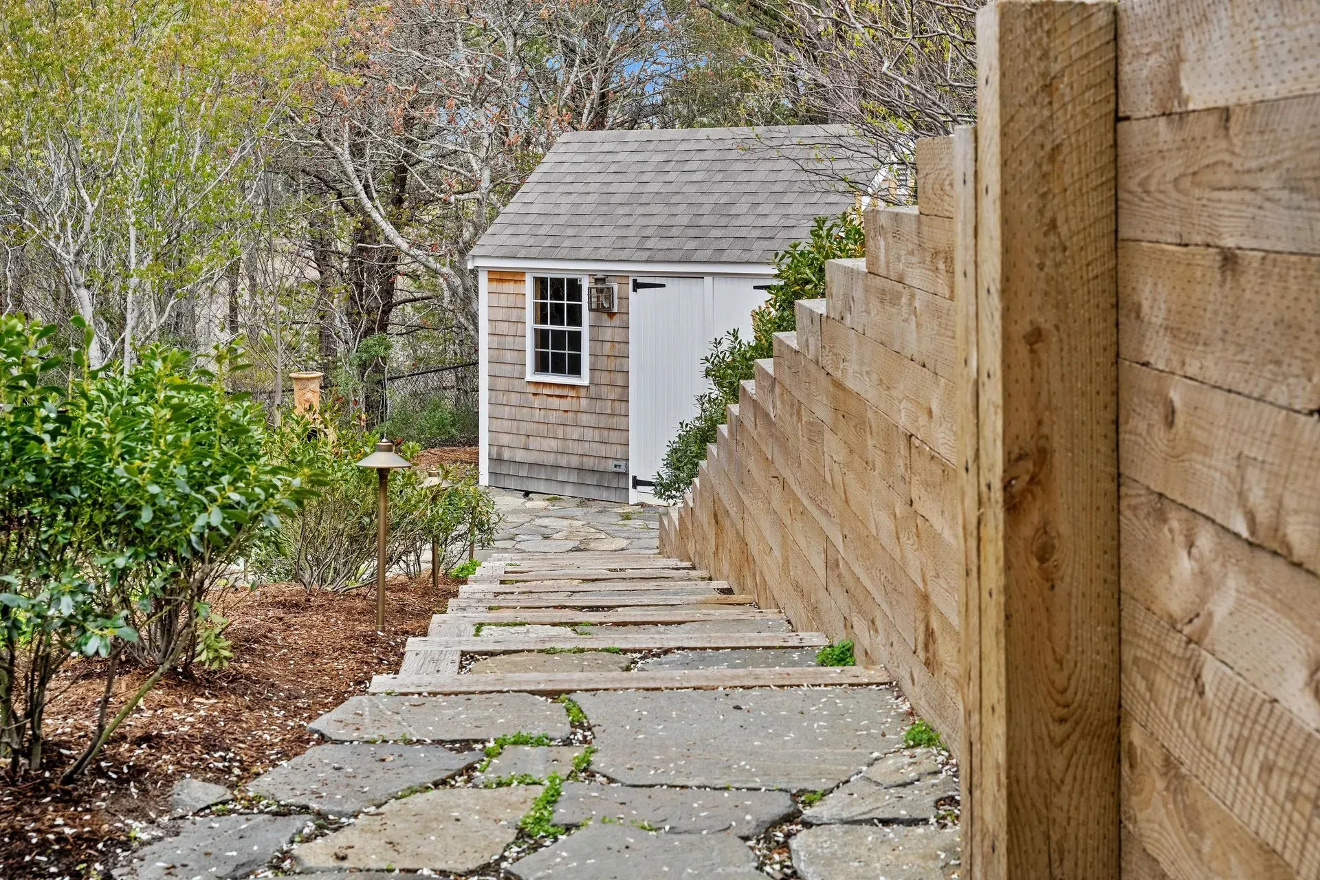 Stone steps beside a wooden fence lead down to a small gray shed in a wooded yard.