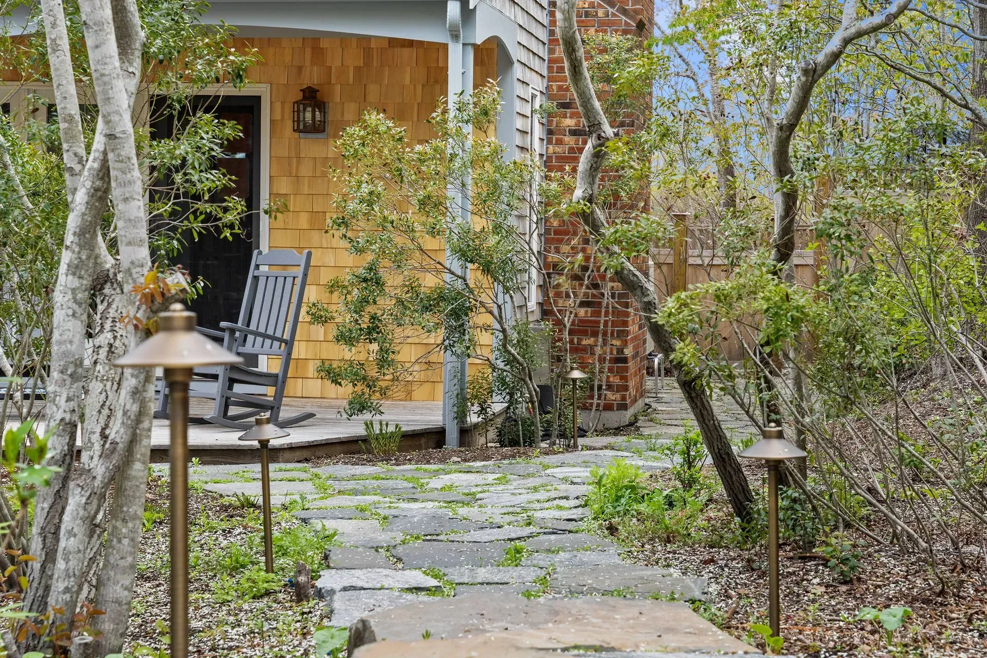 Stone path to a rustic cabin porch with a black rocking chair, lanterns, and birch trees.