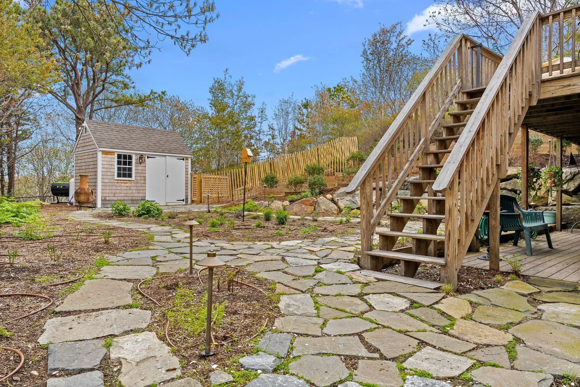 Stone patio and wooden staircase in a backyard garden with a small shed and trees under a blue sky