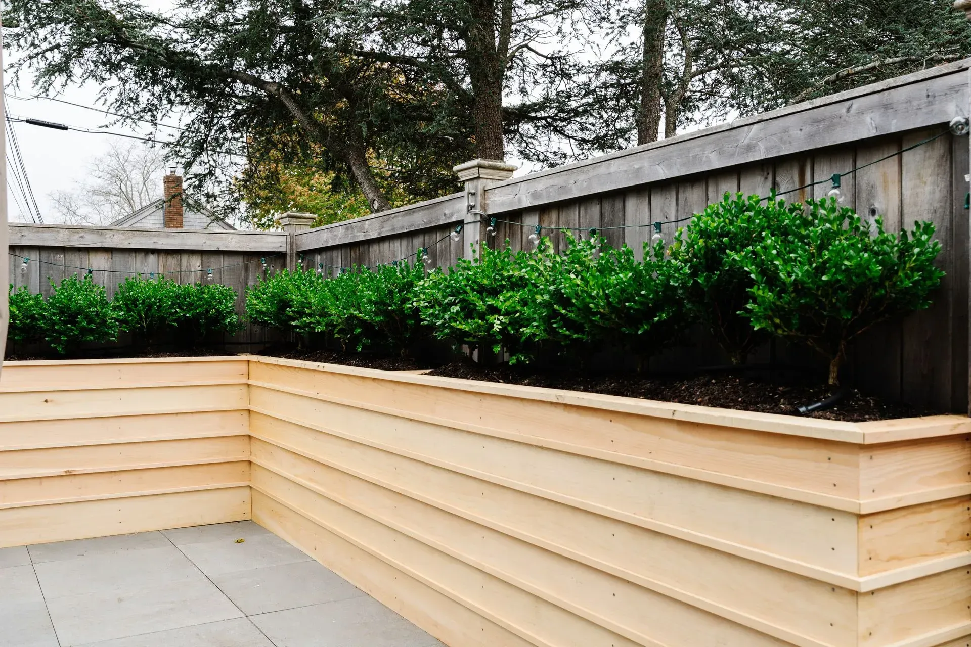 Raised beige planter boxes with green shrubs along a wooden fence on a patio