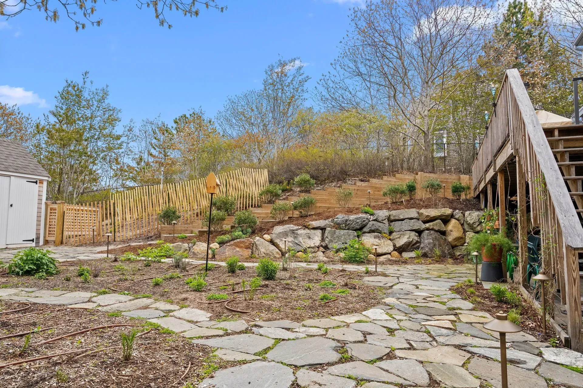 Rocky garden terrace with stone path, wooden stairs, and spring trees under a blue sky