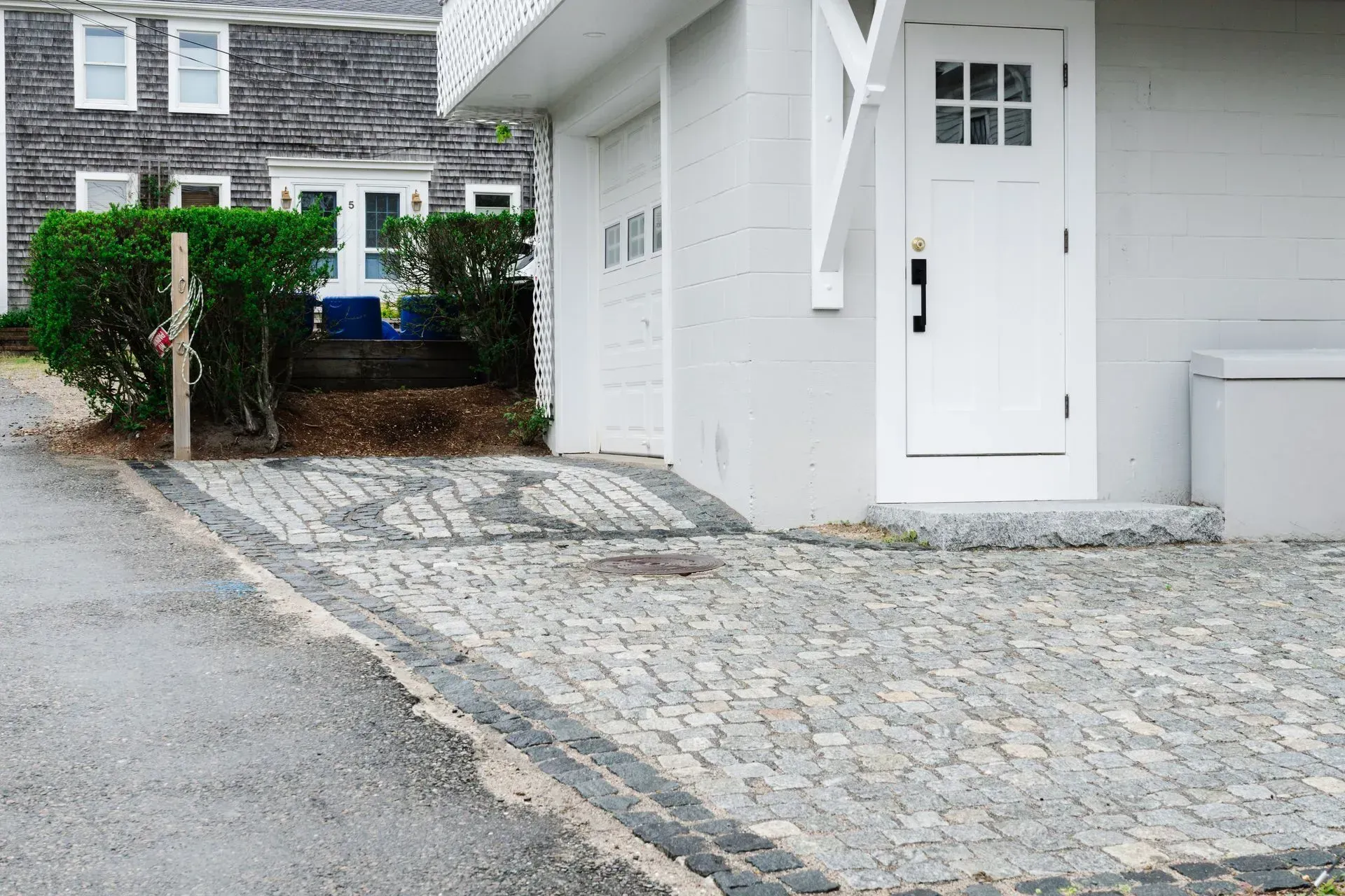 Cobblestone driveway leading to a white house entrance and garage beside shrubbery