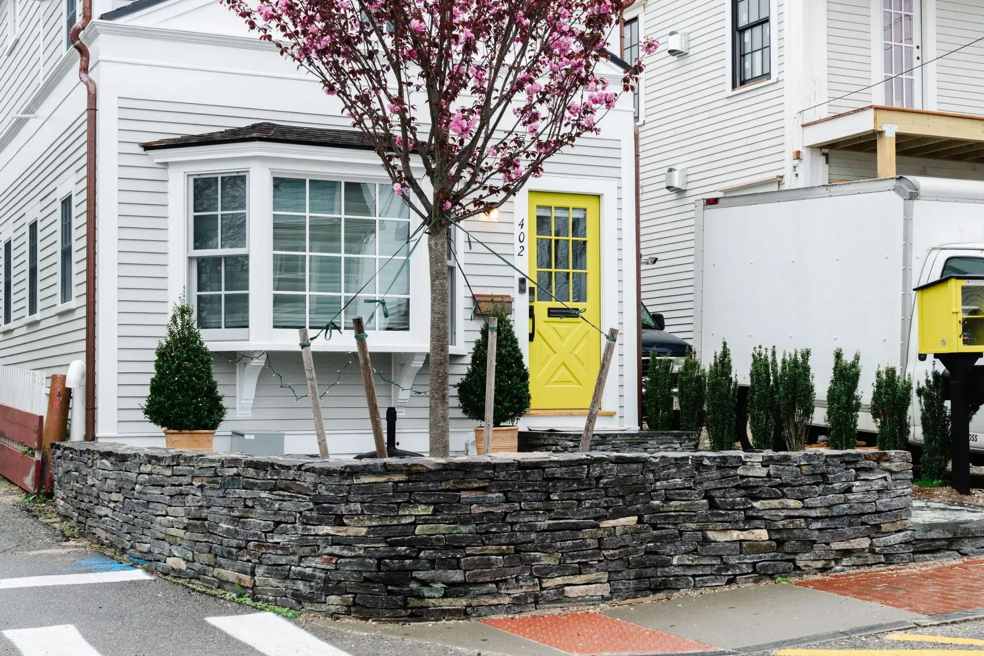 White house with a bright yellow door, bay window, and stone retaining wall under a blooming tree.