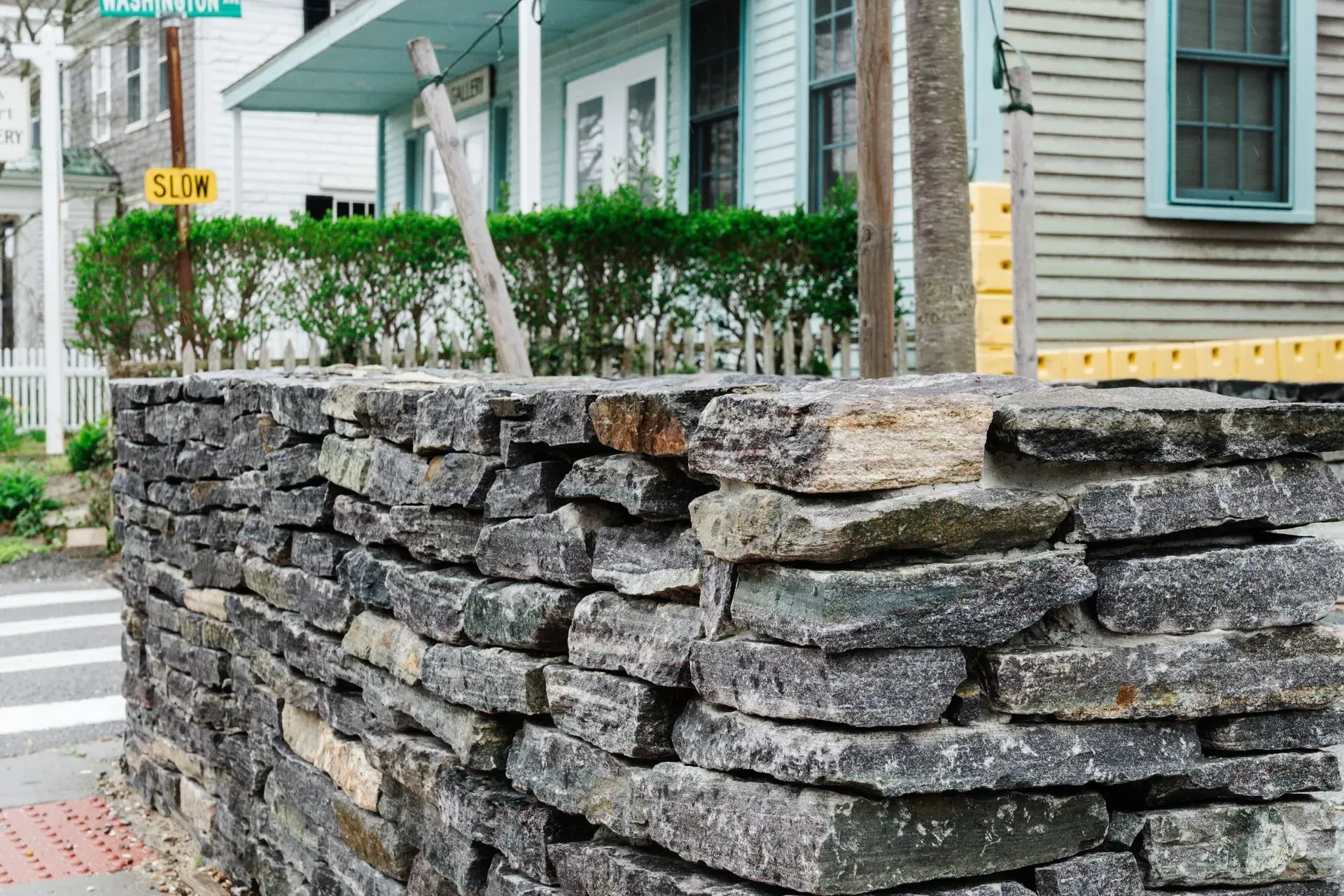 Stone wall beside a yellow house with green shutters and hedge along a sidewalk corner