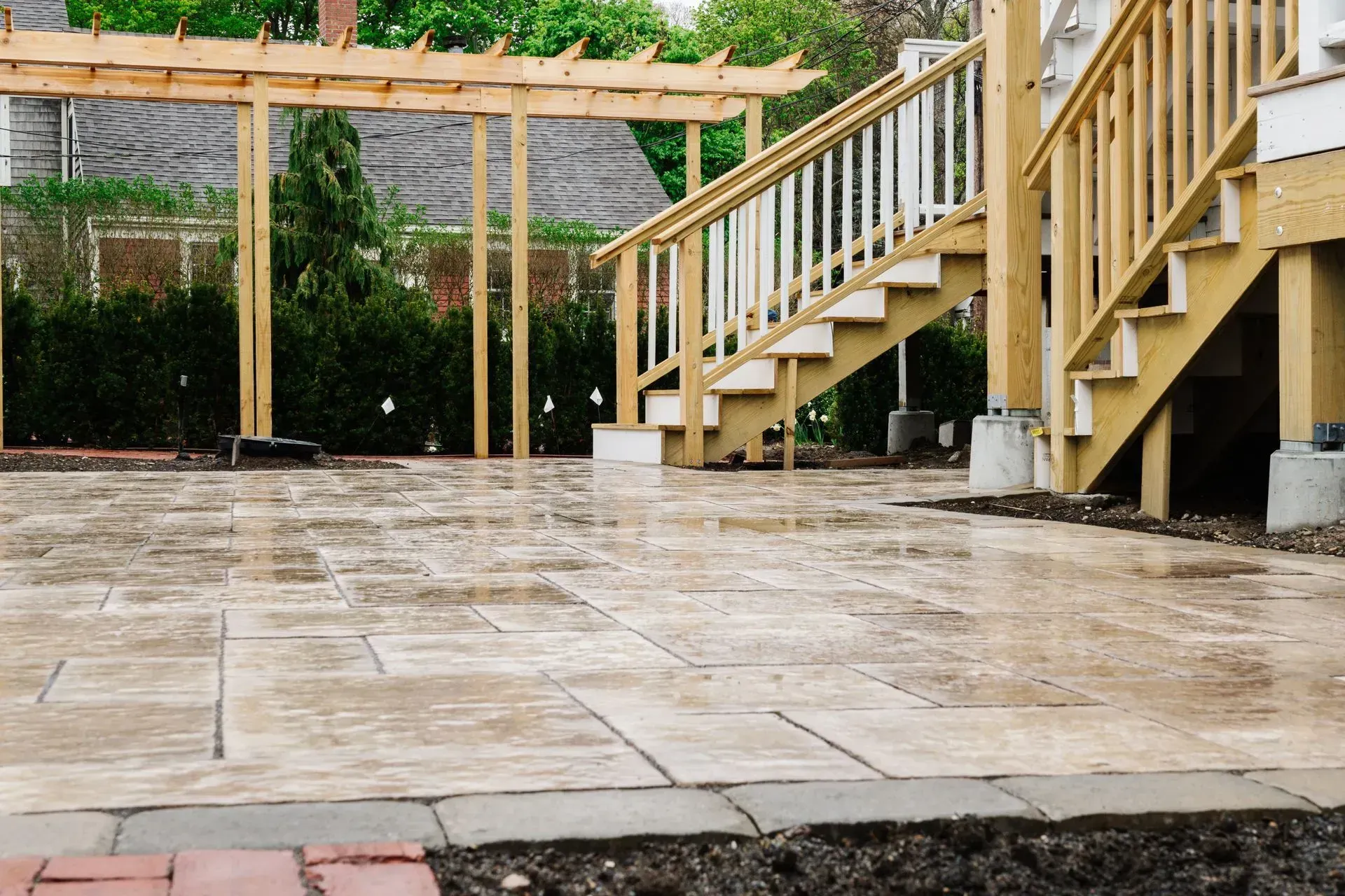 Stone patio beside a wooden deck and stairs in a backyard, with a pergola and privacy fence.