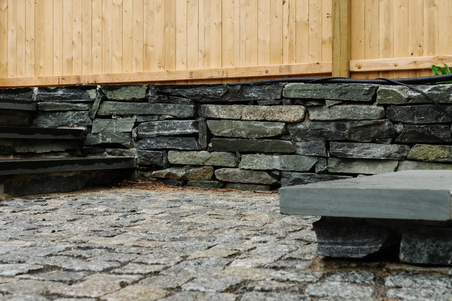Stone-walled patio with wooden fence and a low bench on a cobblestone ground.