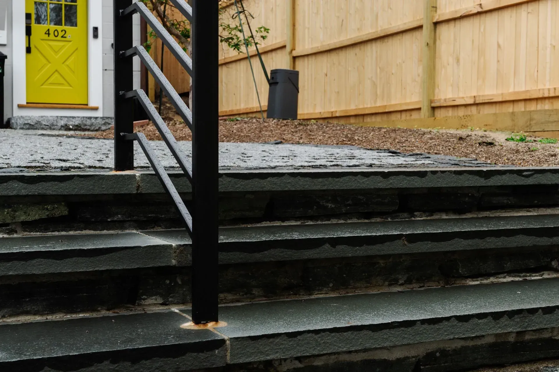 Yellow front door beside dark stairs with black railings and a wooden fence