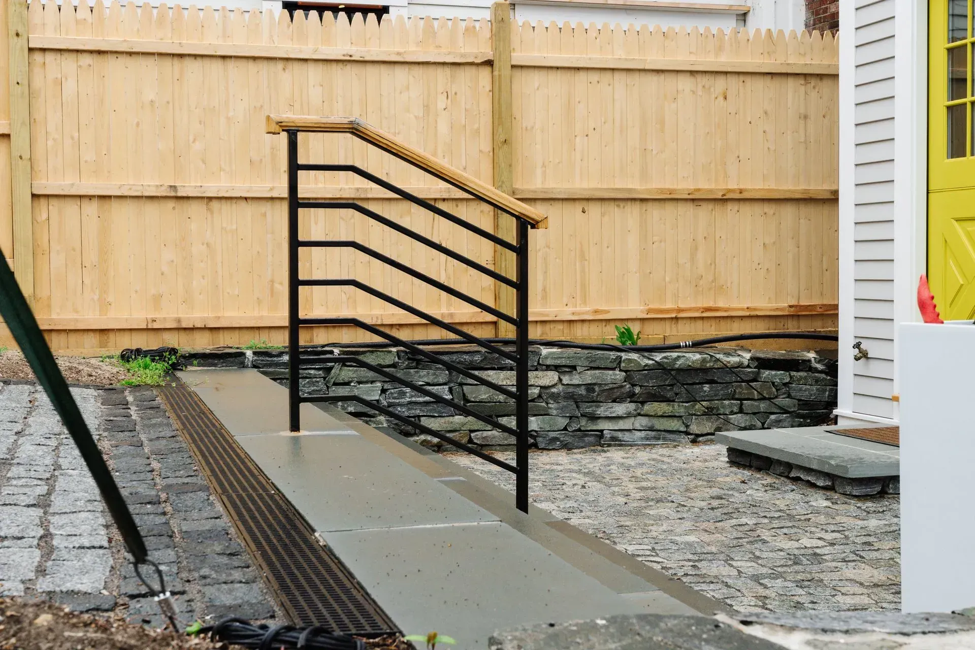 Black metal ramp railing along a concrete path beside a fenced yard and house exterior