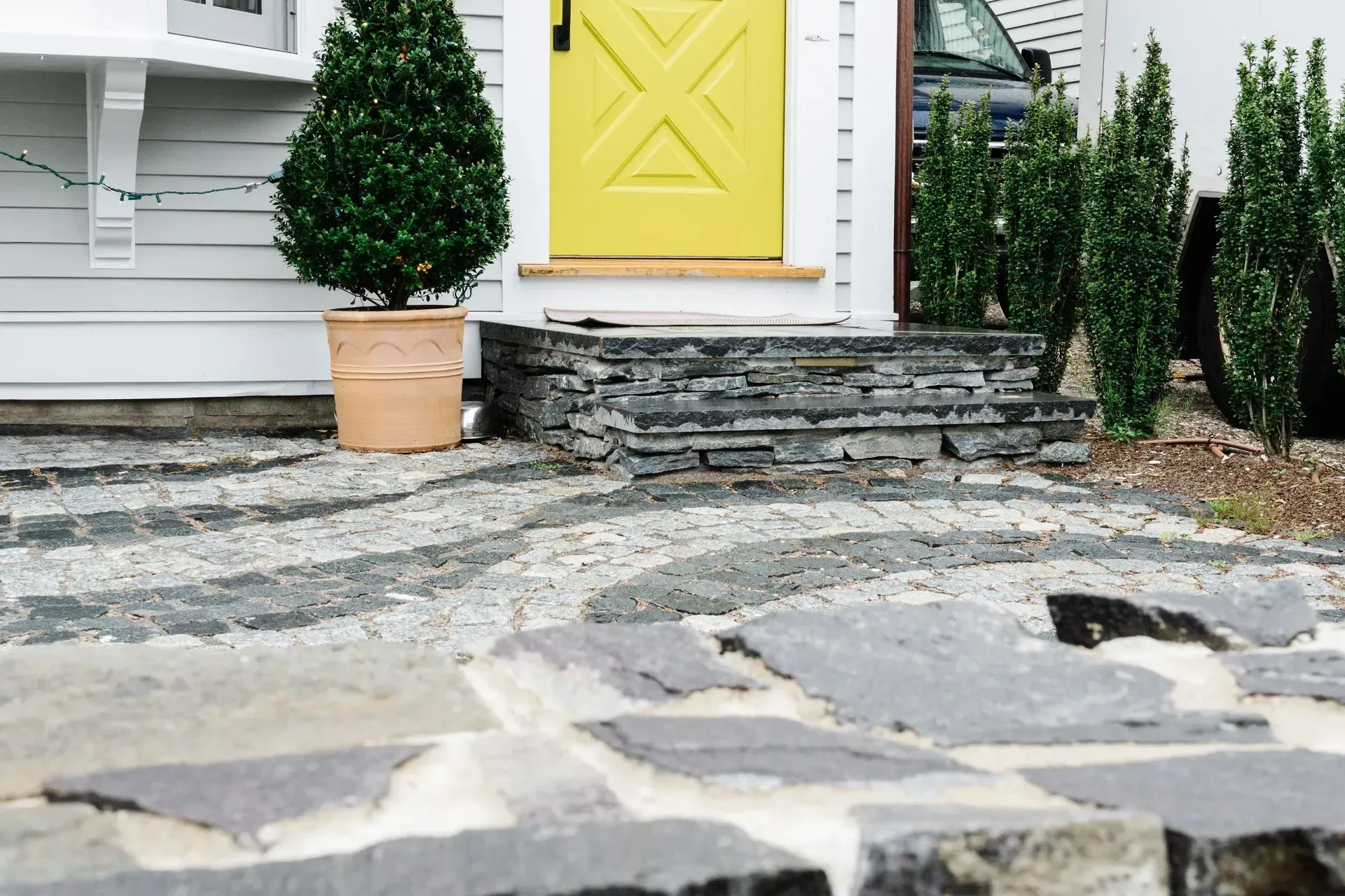Stone walkway leading to a house entrance with yellow door, steps, and potted shrubs