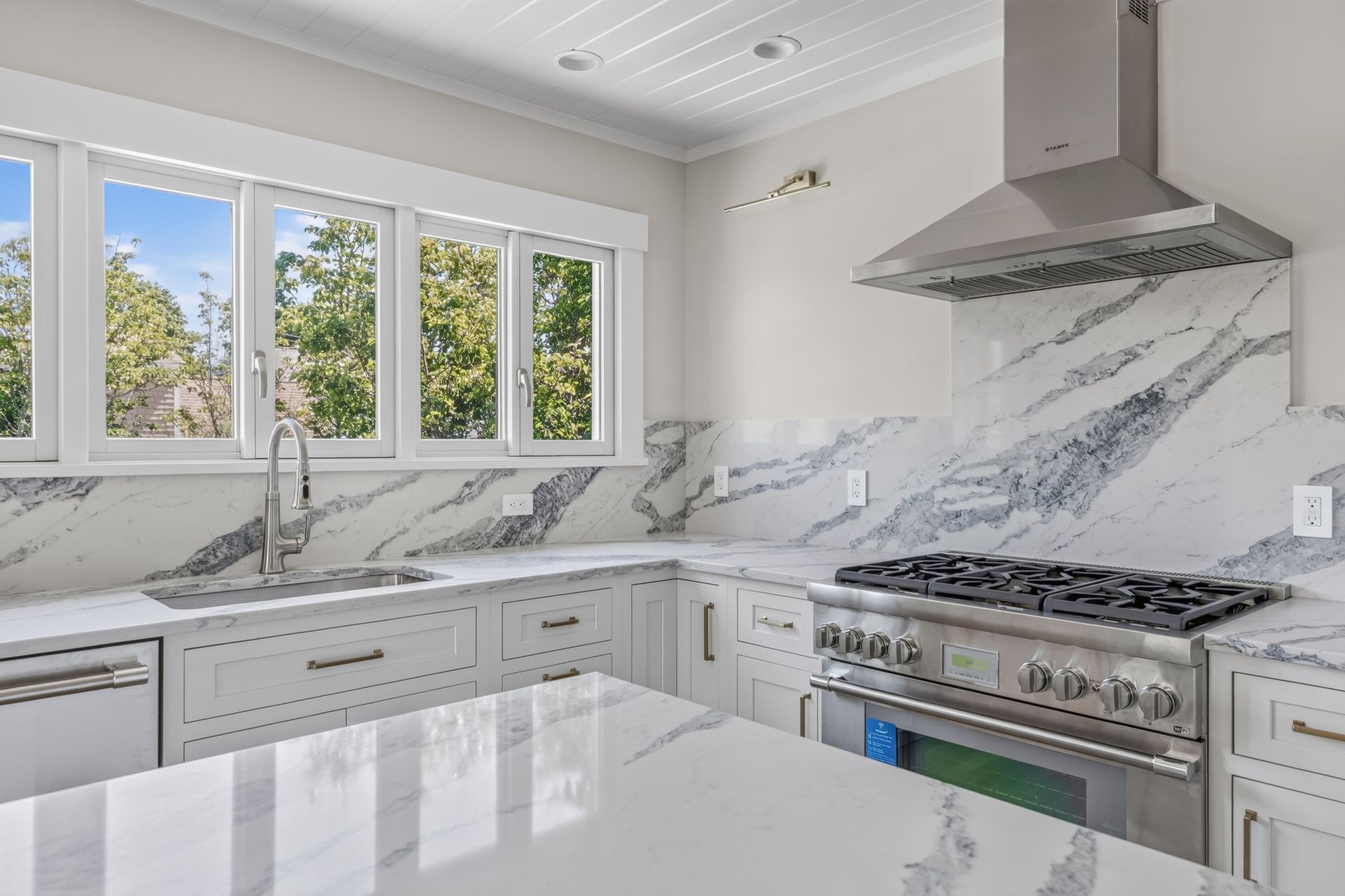 Bright white modern kitchen with marble countertops, large windows, and stainless steel stove and range hood