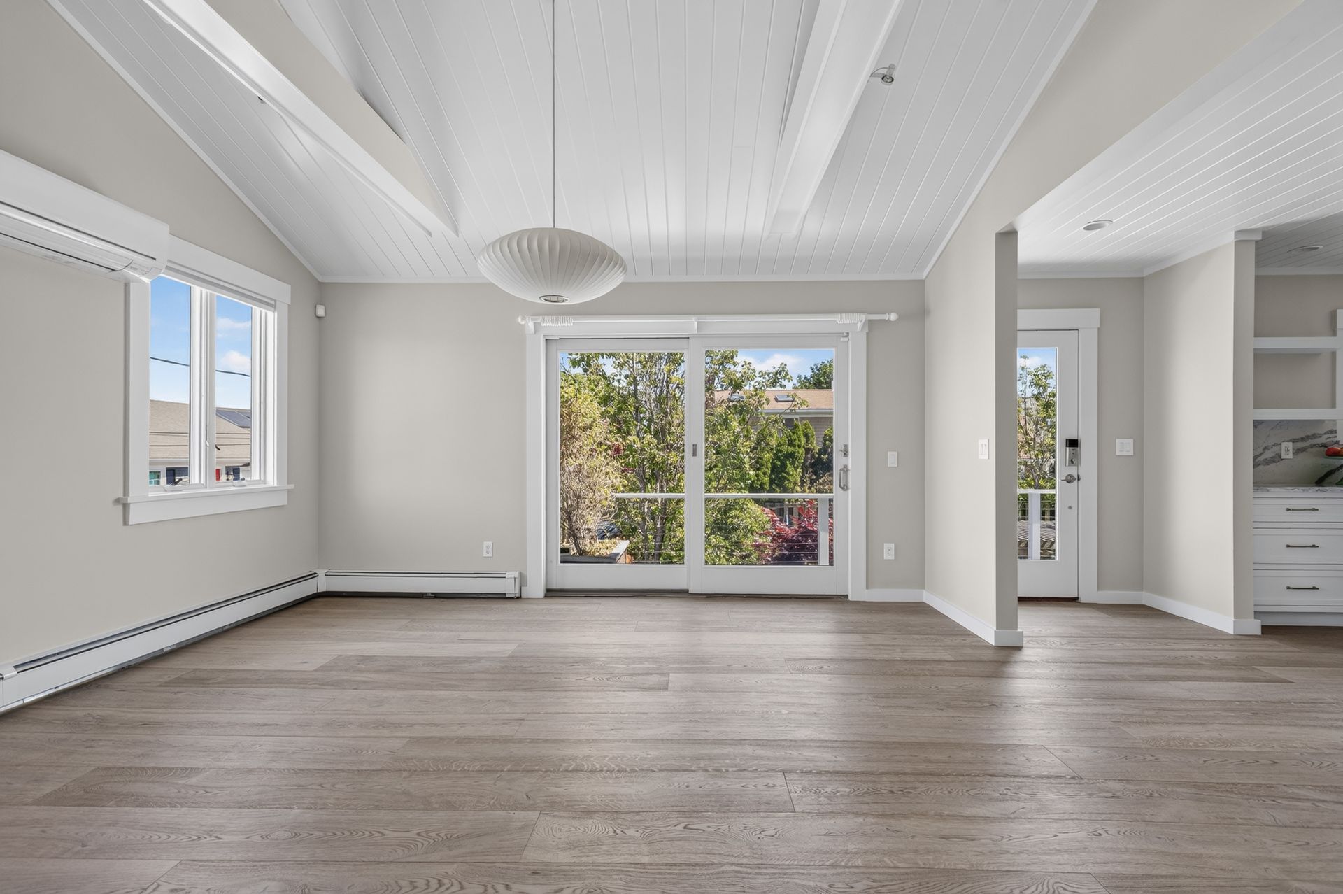 Empty modern living room with white walls, wood floors, large windows, and sliding glass doors to a balcony