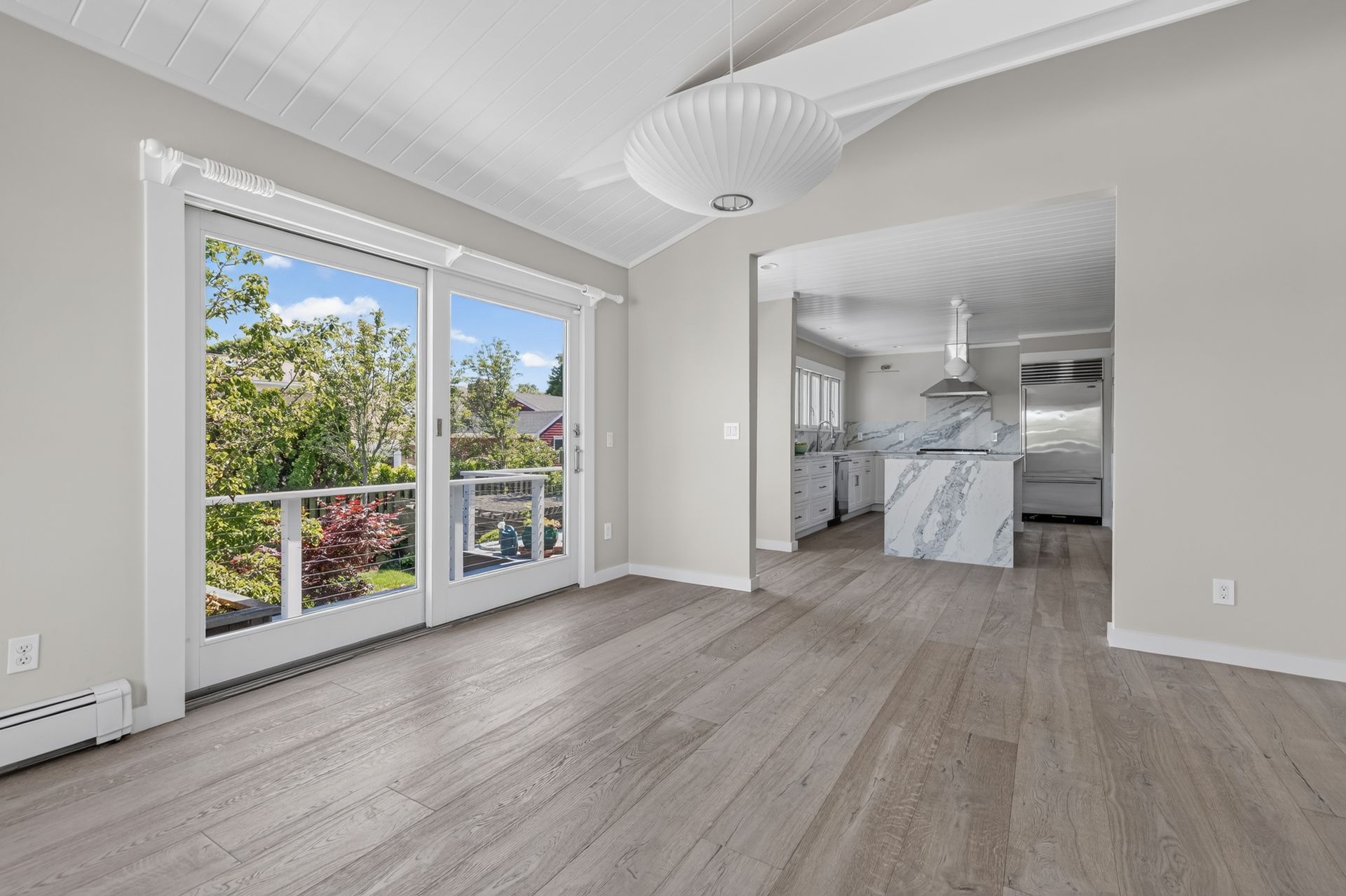 Bright empty living room with large windows, light wood floors, and an open view into the kitchen