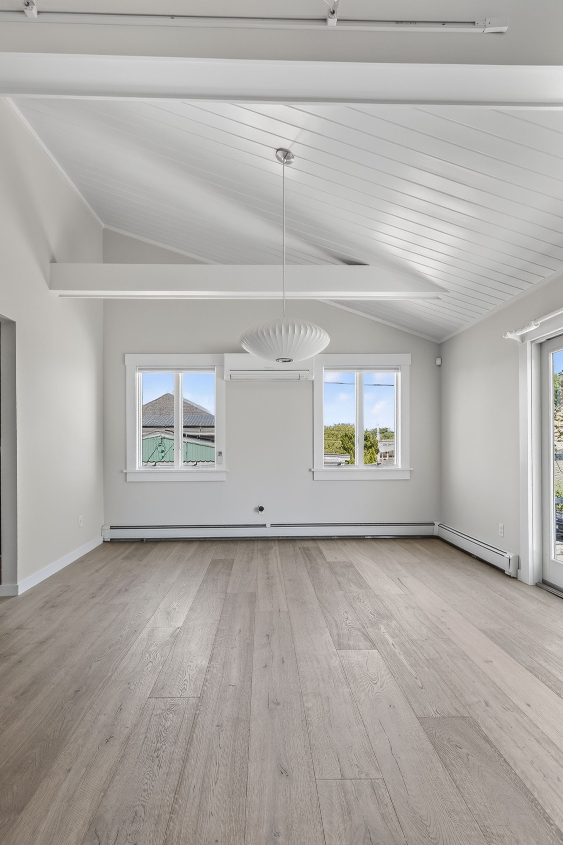 Bright empty living room with white walls, vaulted ceiling, windows, and light wood floors