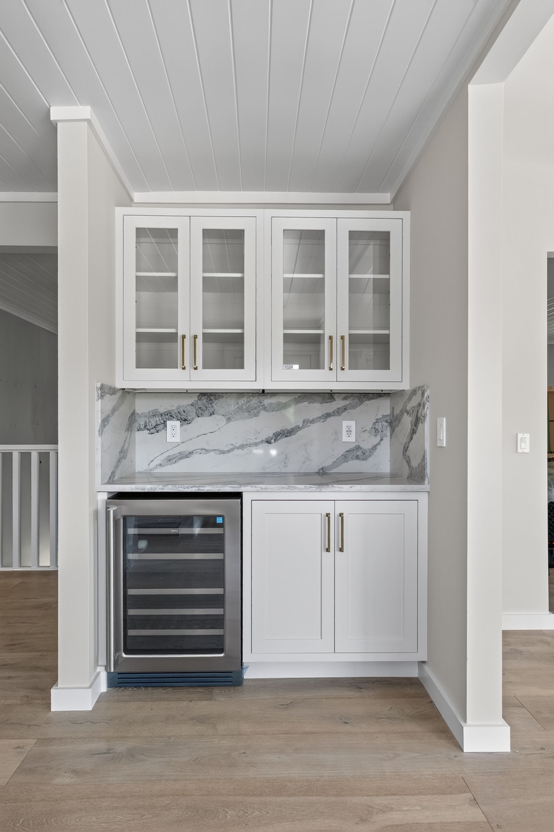 Small built-in kitchenette with glass cabinets, marble backsplash, mini wine fridge, and white cabinetry.