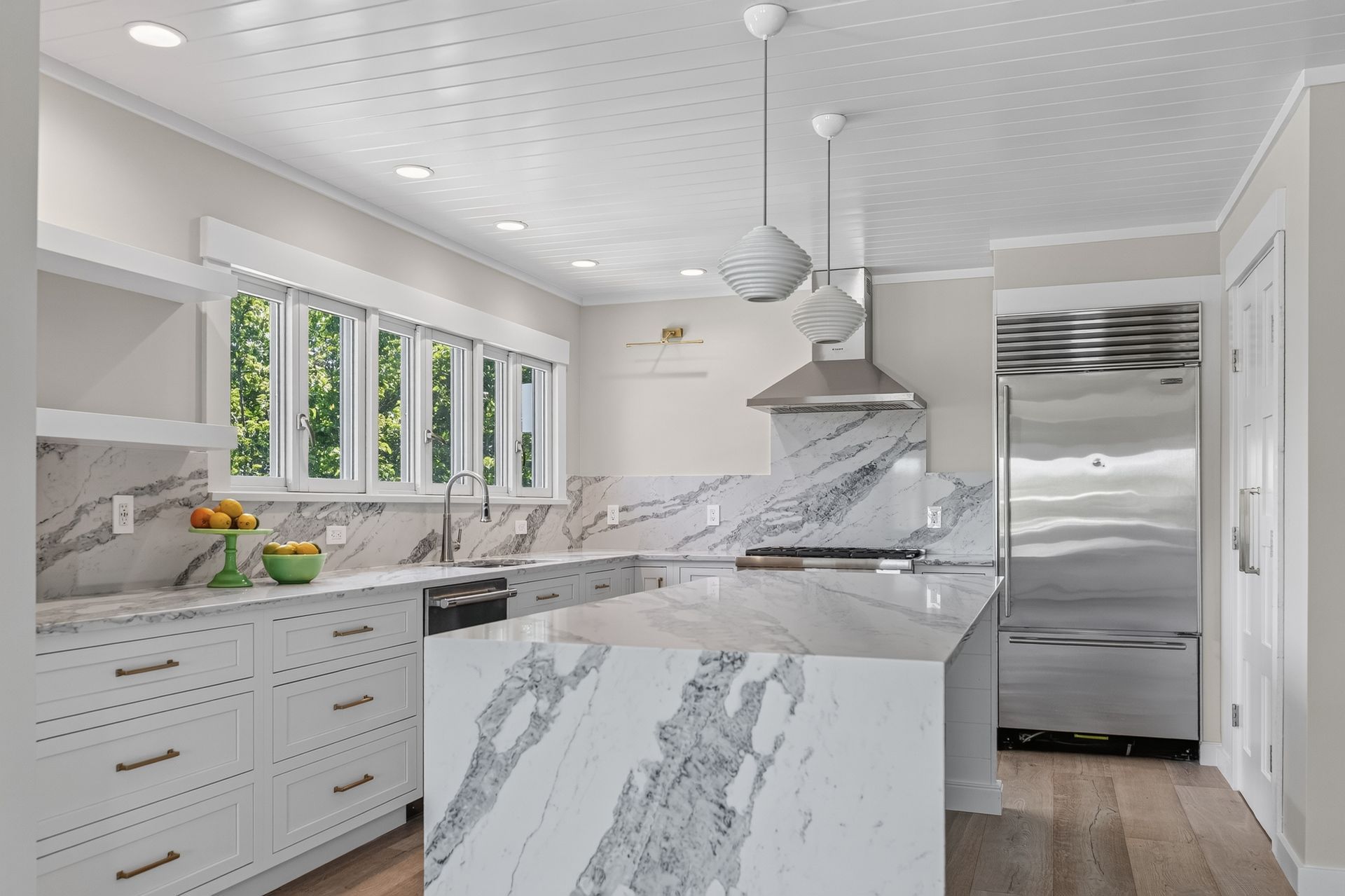 Bright white modern kitchen with marble island, stainless fridge, and large windows.