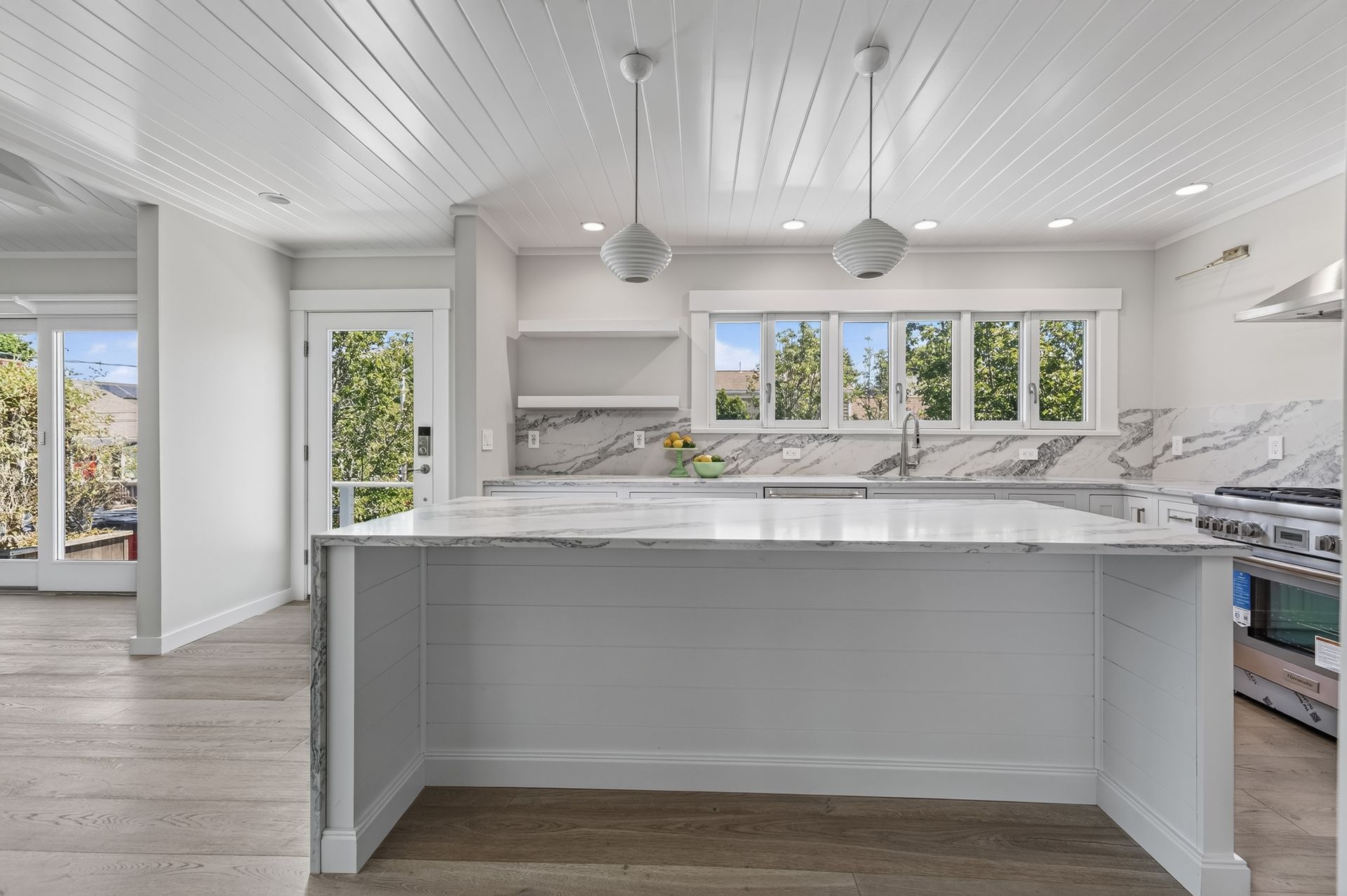 Bright modern kitchen with white cabinets, marble island, and large windows overlooking greenery