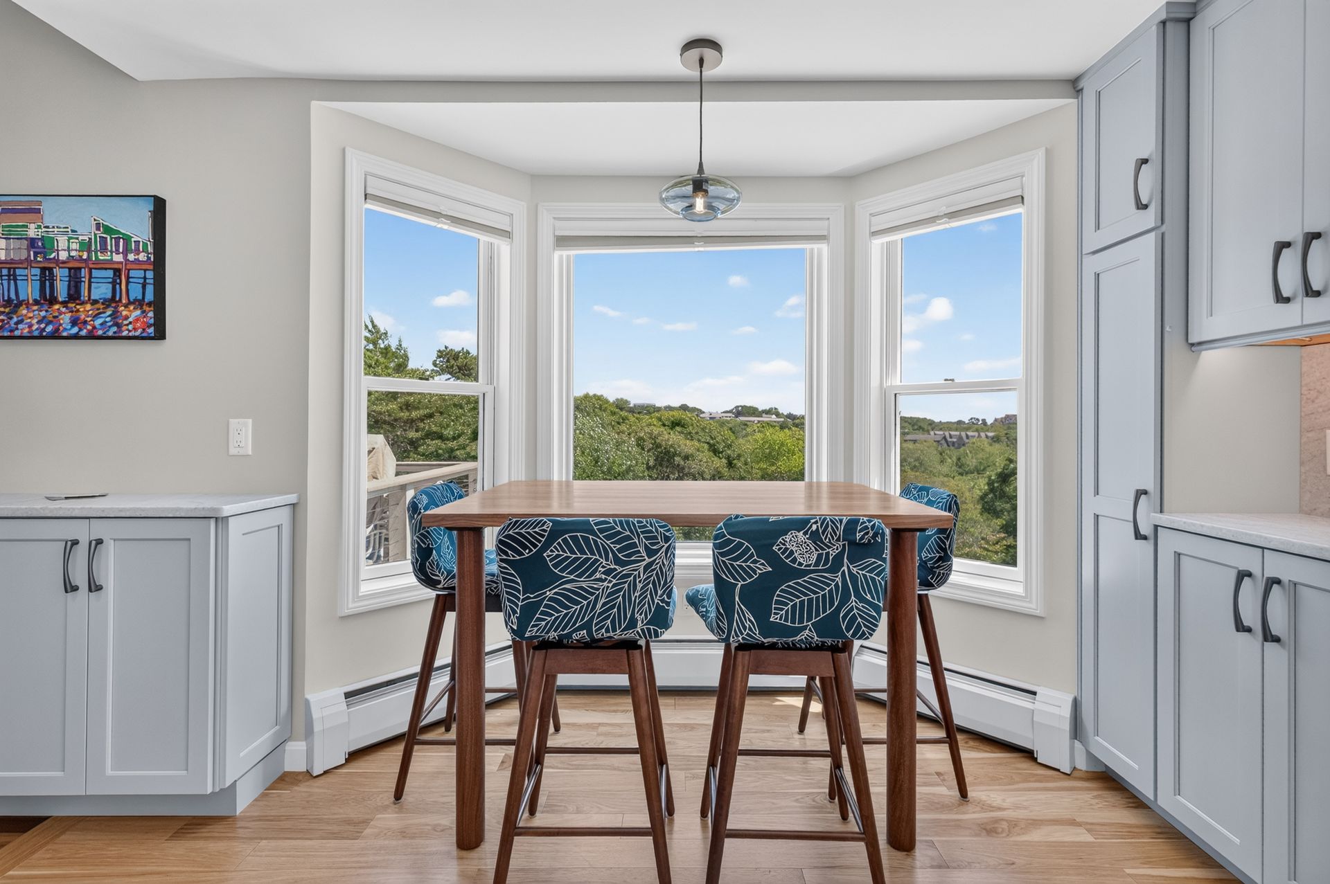 Bright kitchen nook with a wooden table, blue chairs, bay windows, and a view of green trees outside