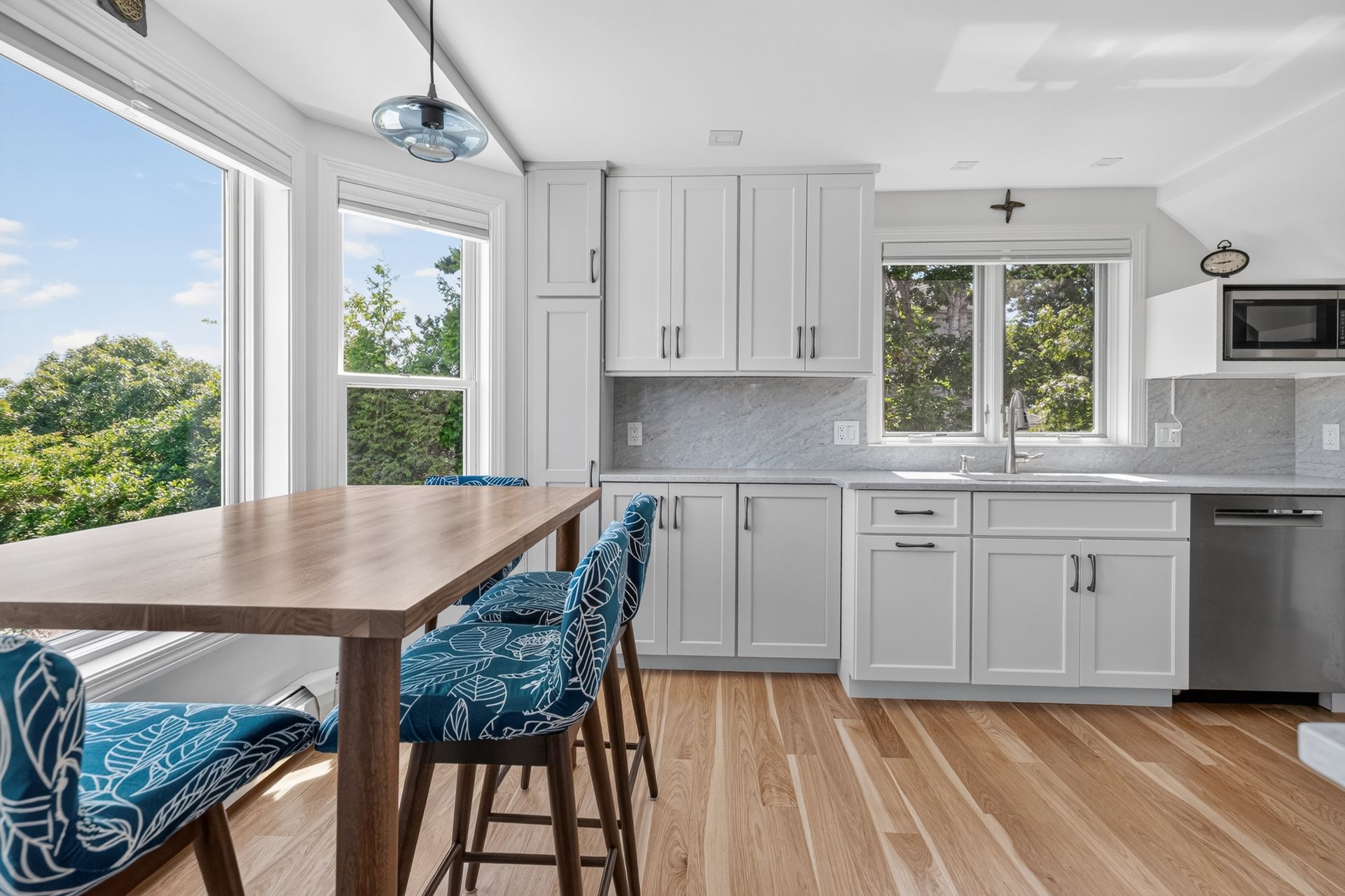 Bright modern kitchen with white cabinets, stainless appliances, wood table, blue chairs, and large windows