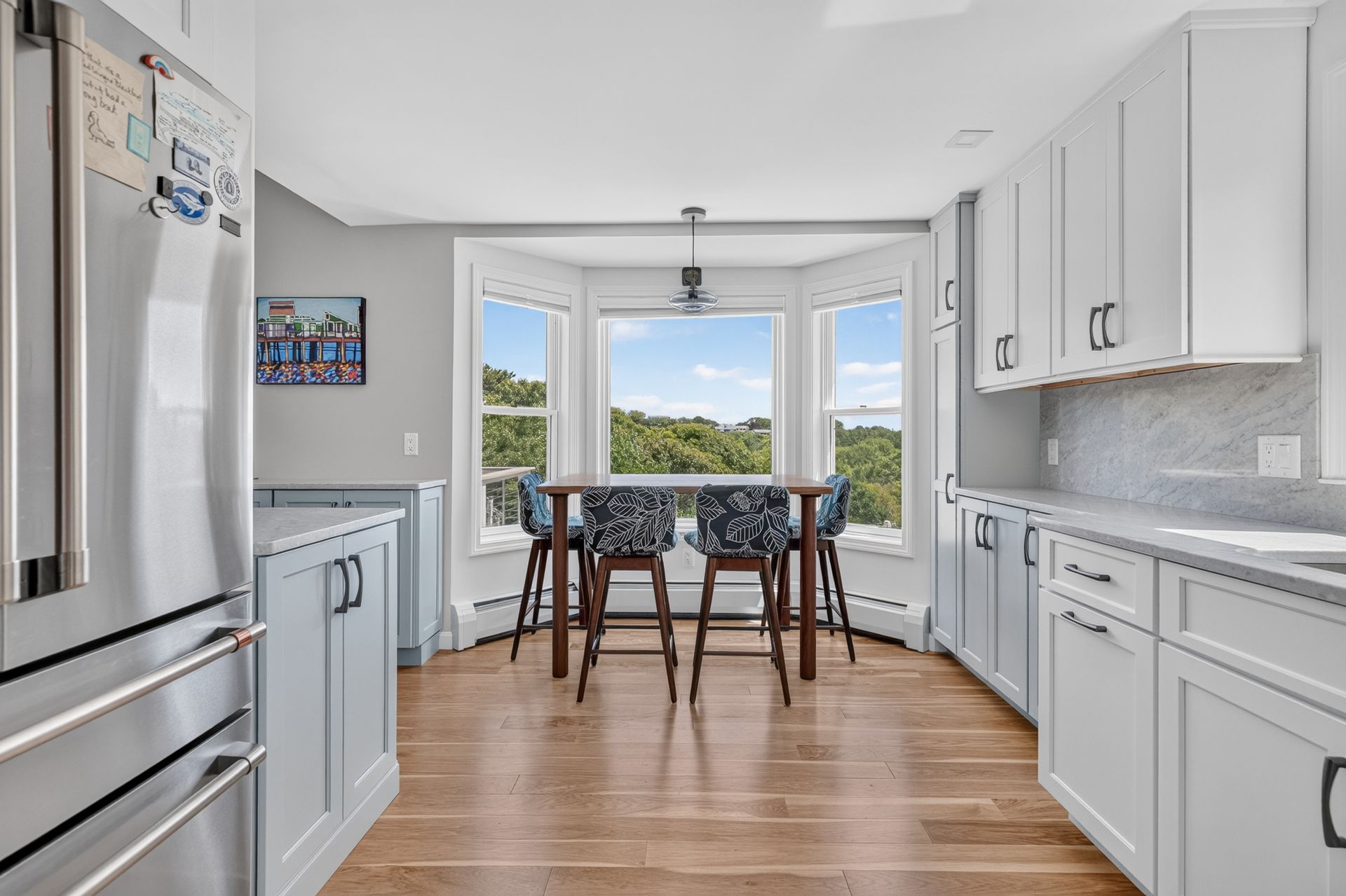 Bright modern kitchen with white cabinets, stainless fridge, wood floor, and dining table by a large window.
