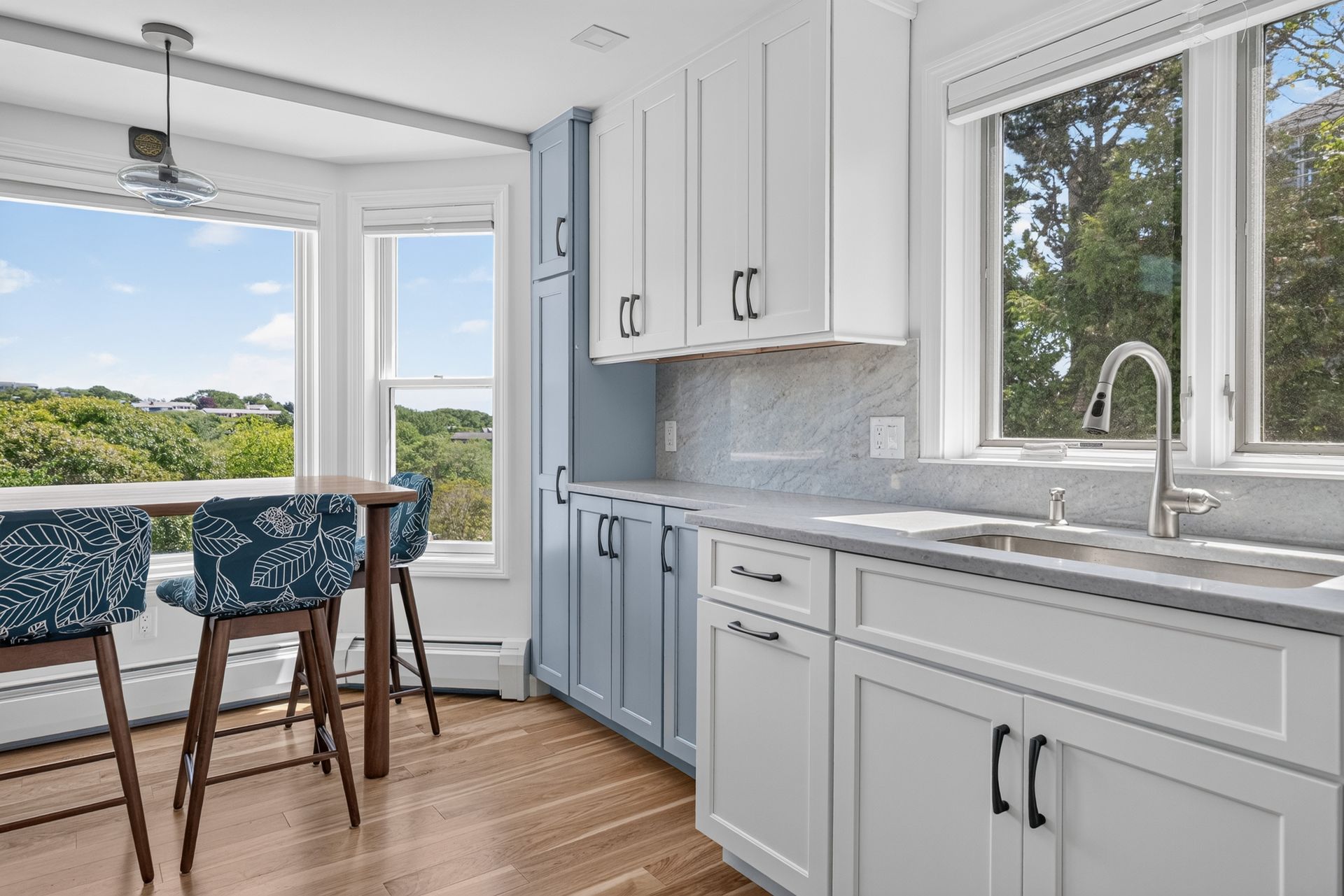 Bright kitchen with white cabinets, blue accents, large windows, and a dining nook overlooking greenery