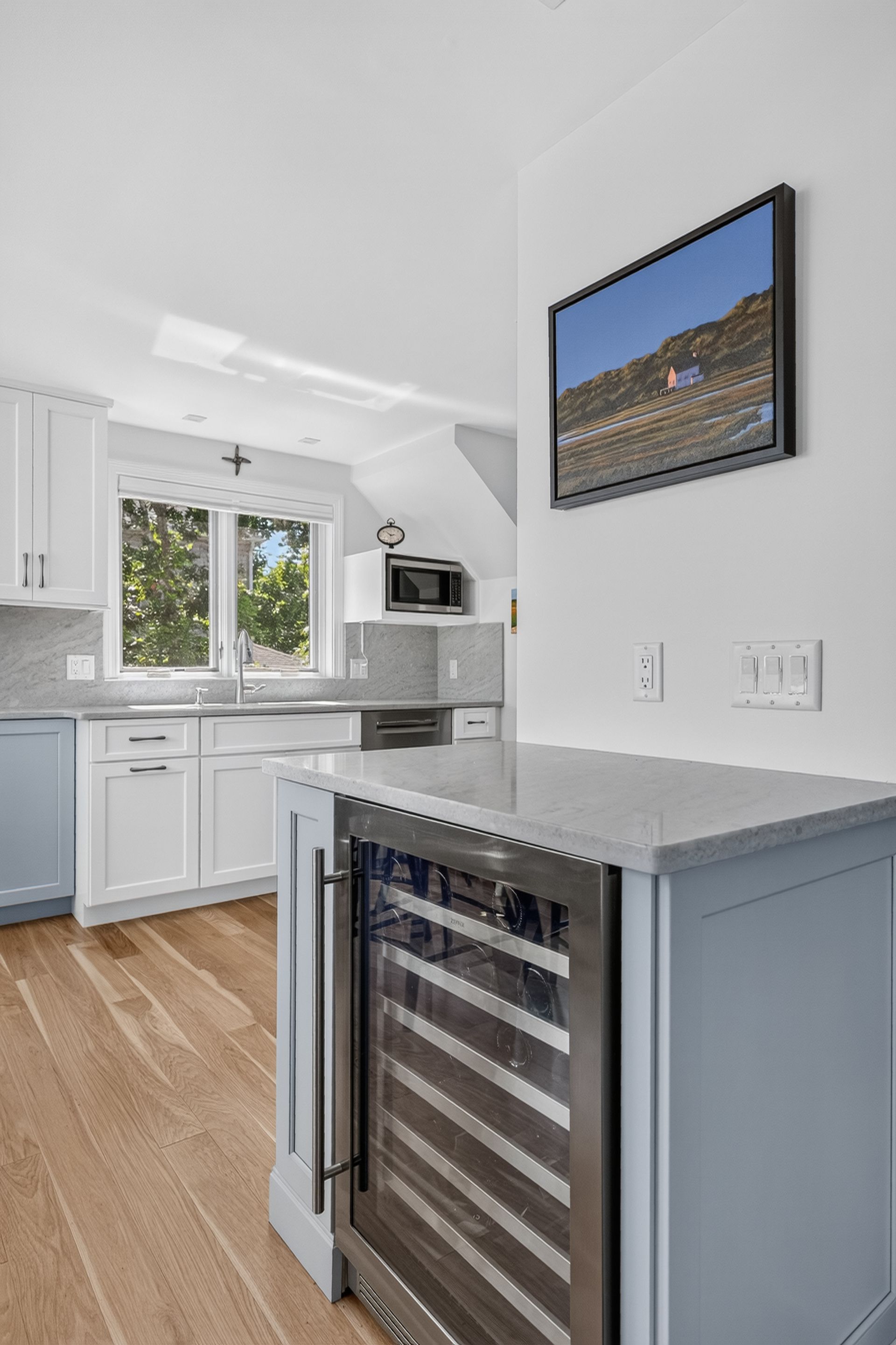 Bright kitchen with white cabinets, gray island, wood floor, and a wine fridge under a framed wall photo.
