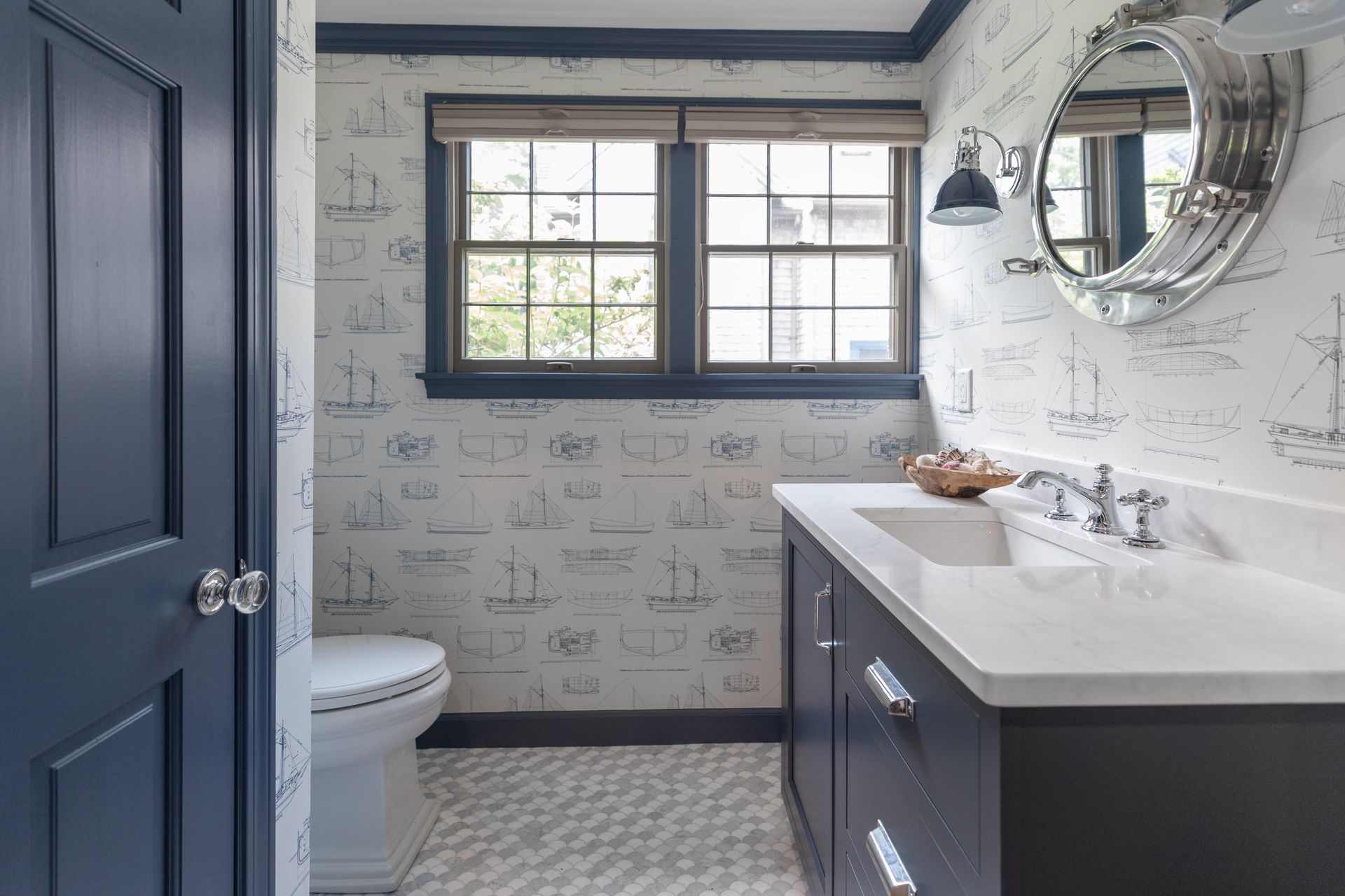 Small bathroom with navy cabinets, white vanity, patterned wallpaper, and a round mirror.