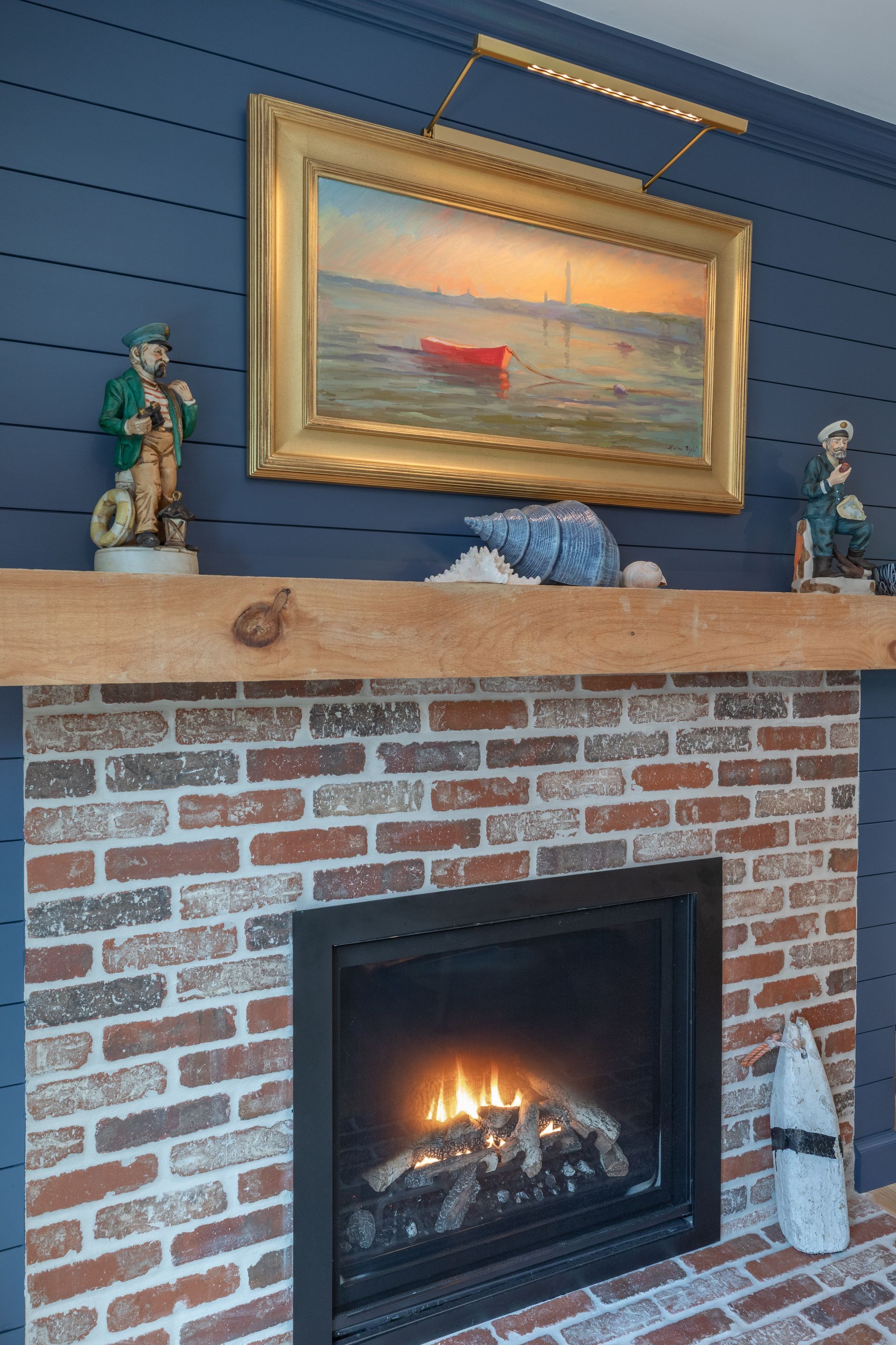 Brick fireplace with lit gas insert, wood mantel, blue wall, gold-framed seascape, and decorative figurines
