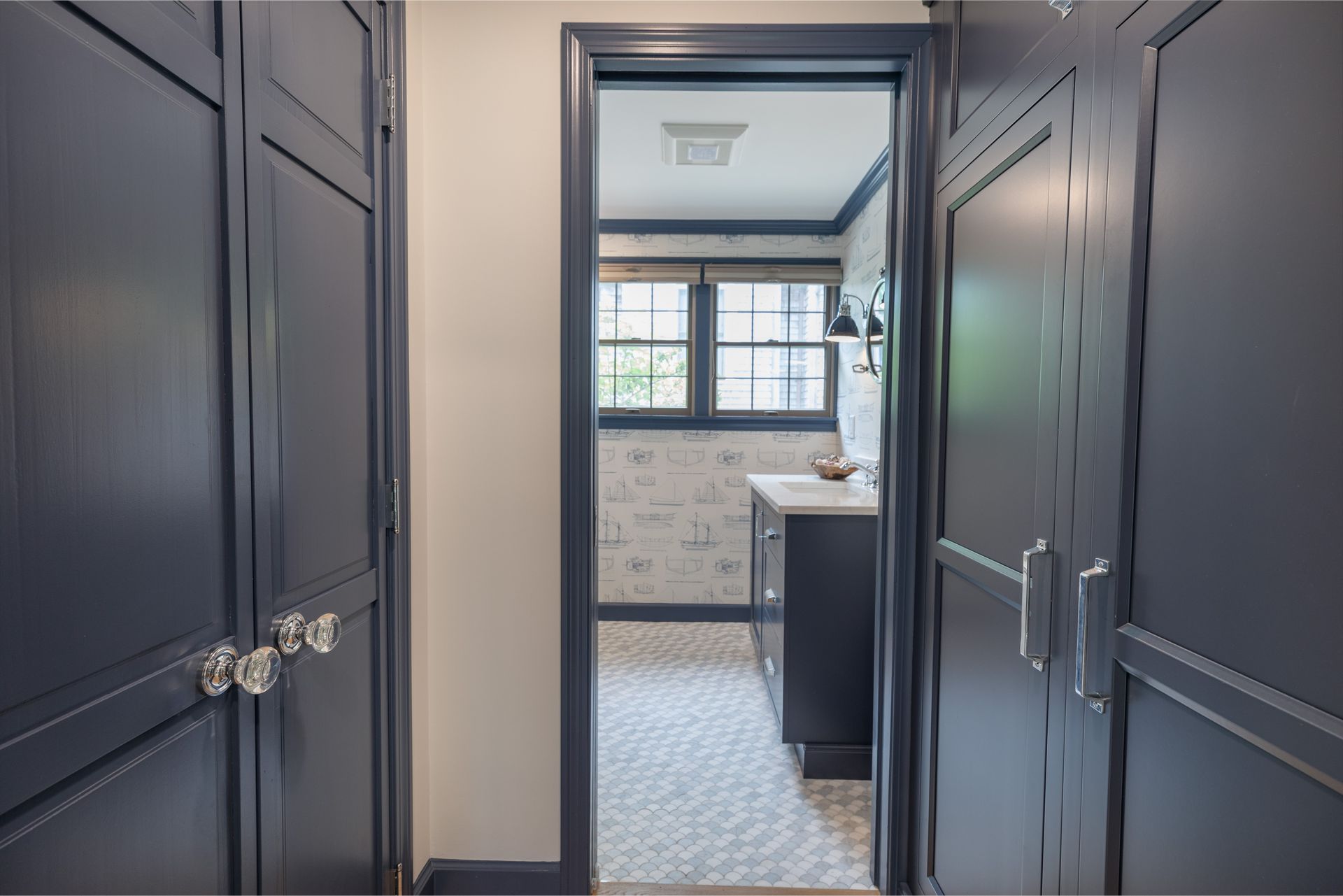 Dark hallway opening into a bright bathroom with blue-gray cabinetry and patterned tile floor