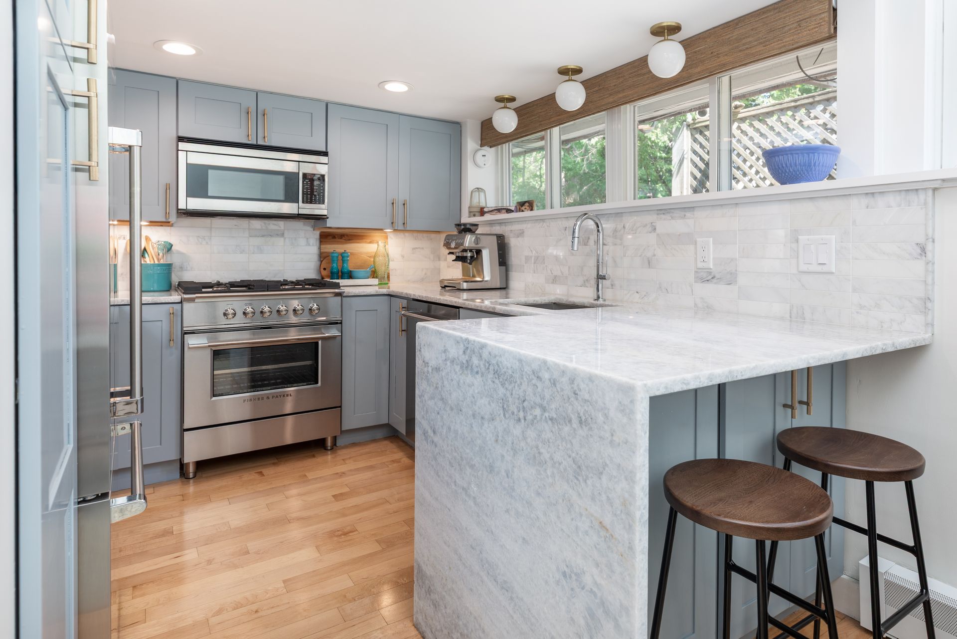 Bright modern kitchen with gray cabinets, marble counters, stainless appliances, and bar stools by a peninsula.