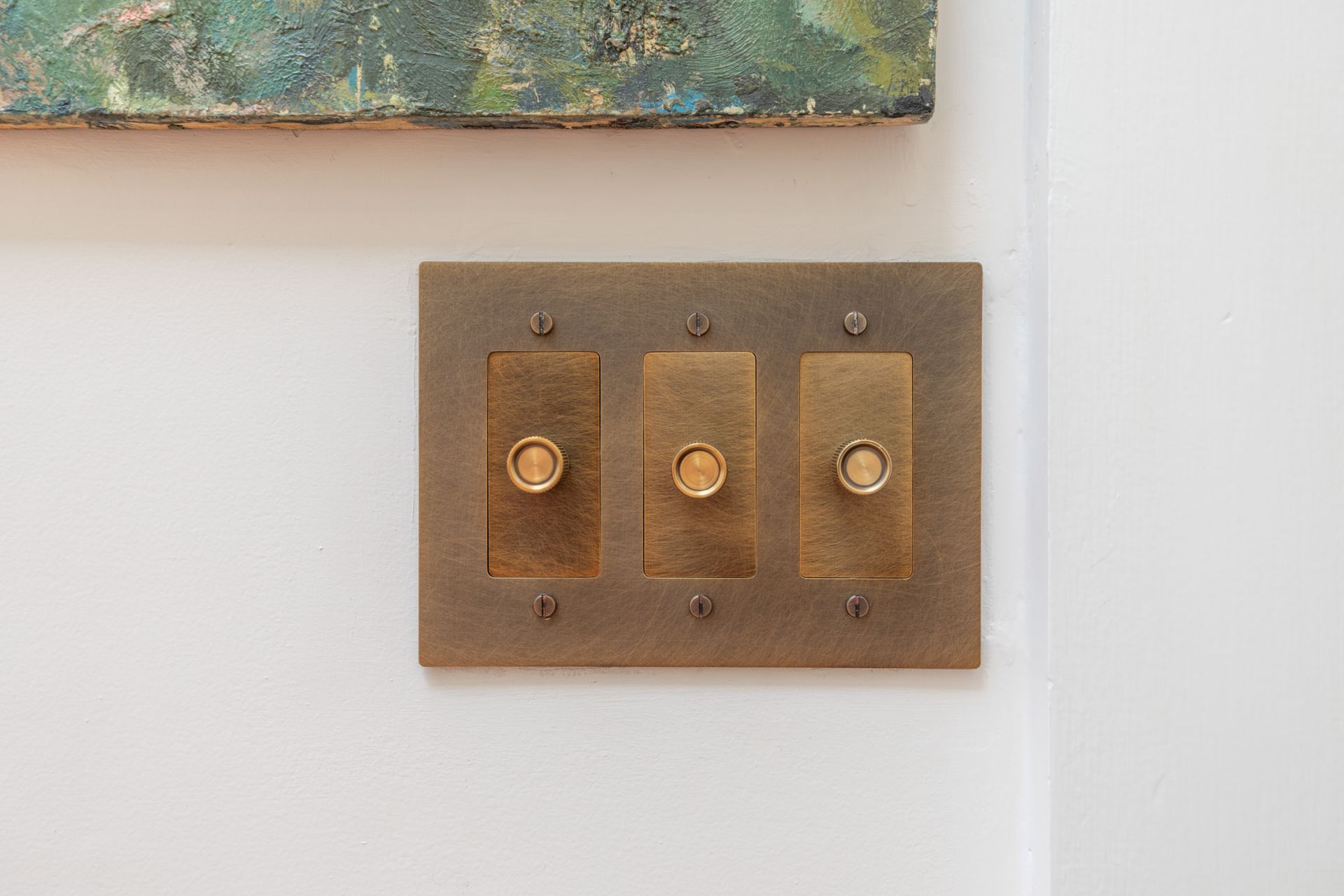 Three brass light switches on a white wall beside framed artwork.