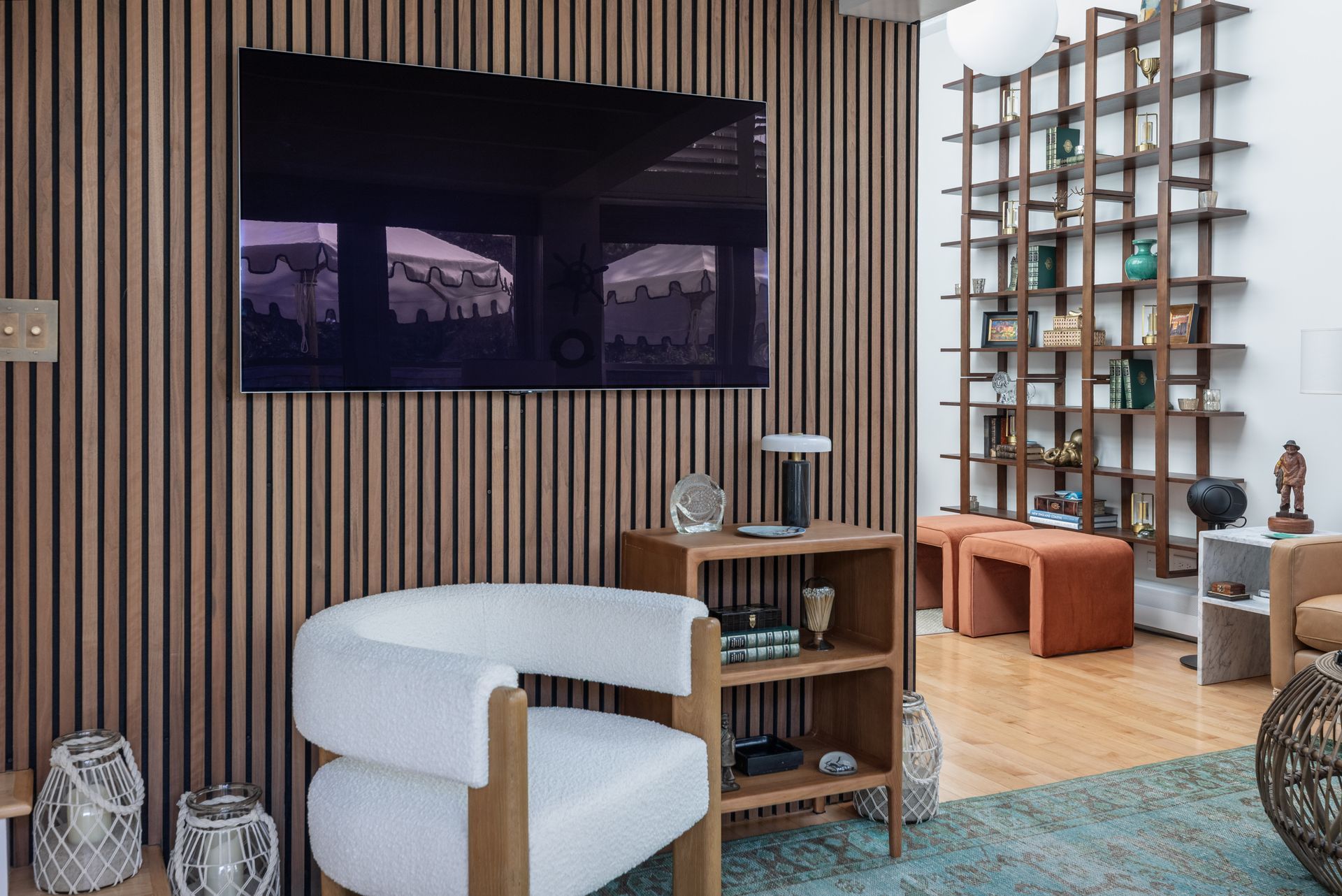 Modern lounge with wood slat wall, large TV, white chair, small table, and shelving beside orange stools.