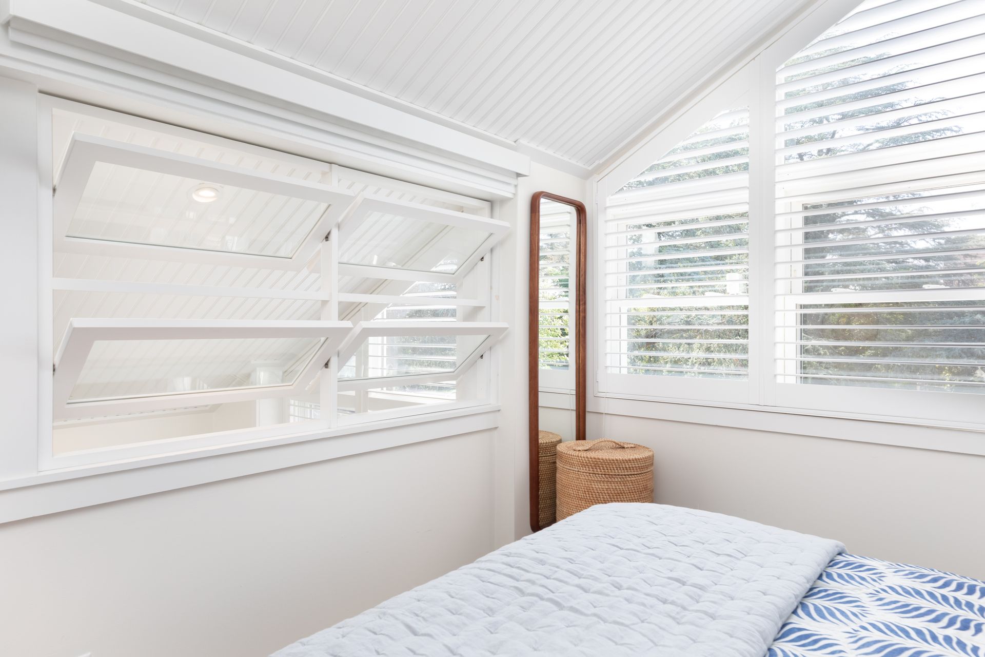 Bright white bedroom with louvered windows, blue bedding, and a wicker basket by the wall