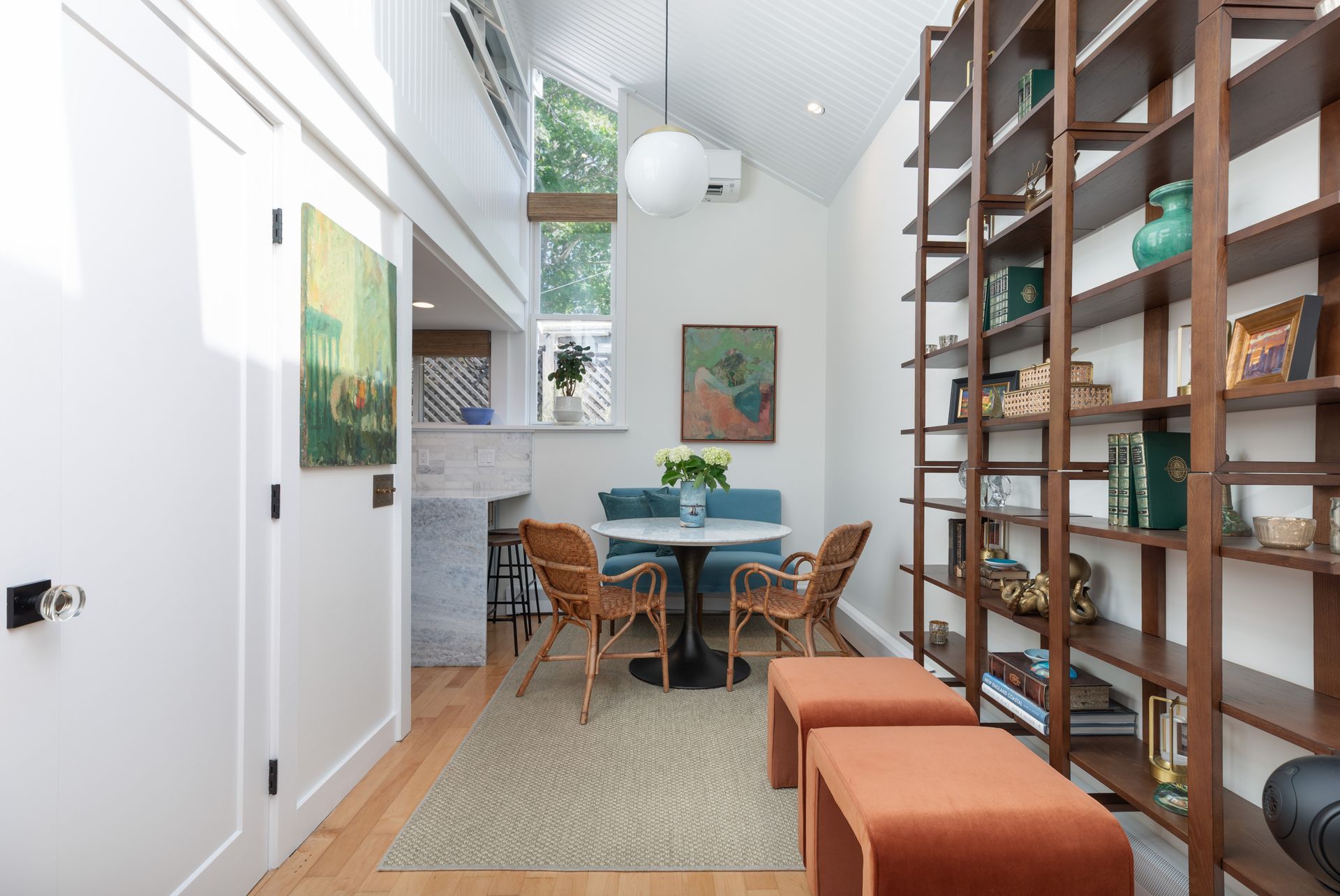 Bright modern dining nook with wooden table, rattan chairs, tall shelves, and orange ottomans under a skylight