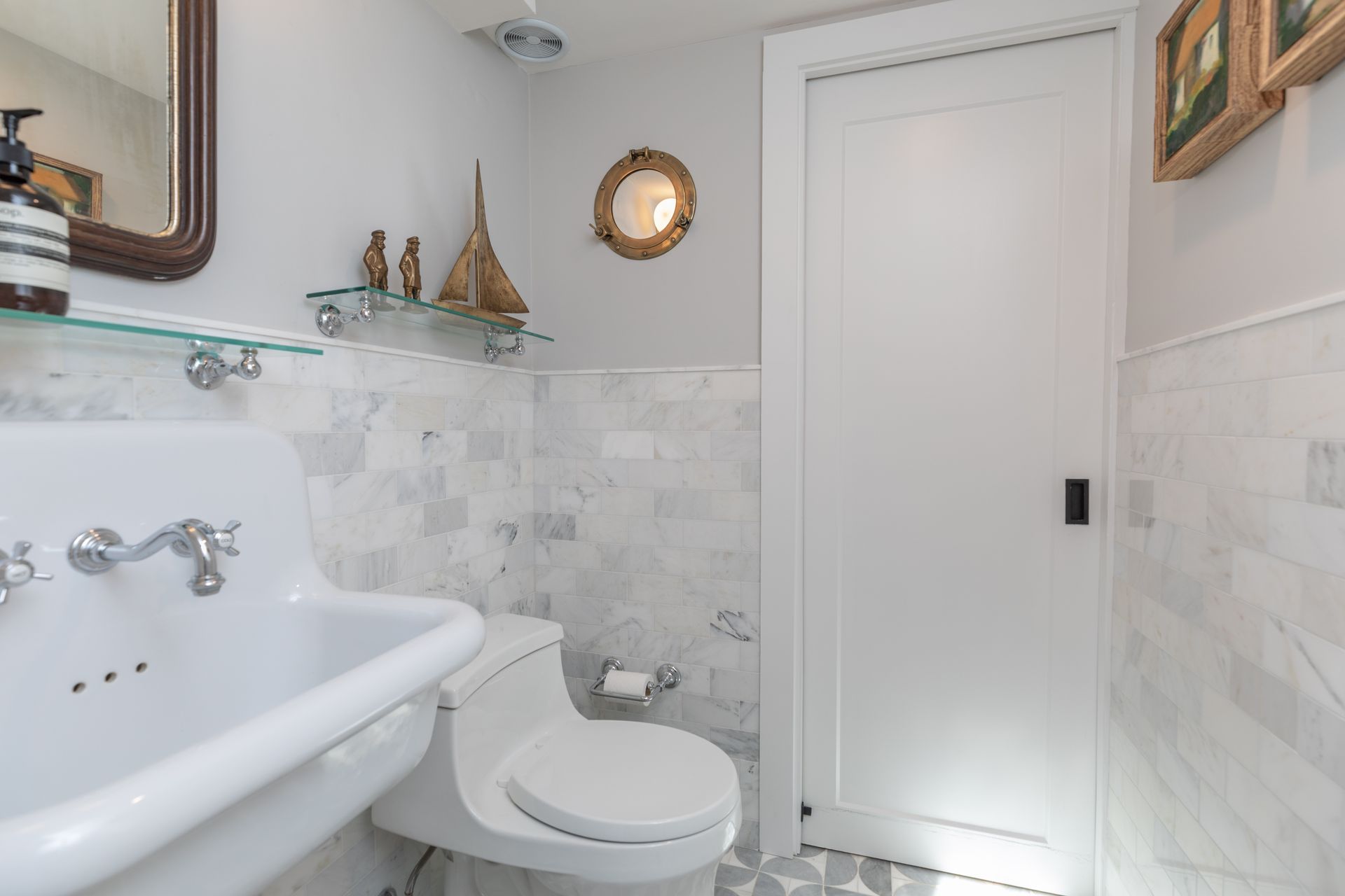 Small white bathroom with marble tile, pedestal sink, toilet, wall shelf, and closed door
