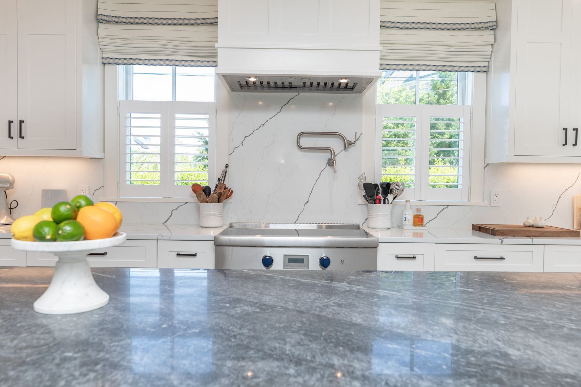 Bright white kitchen with blue-gray countertop, sink, windows, and fruit bowl on a pedestal.