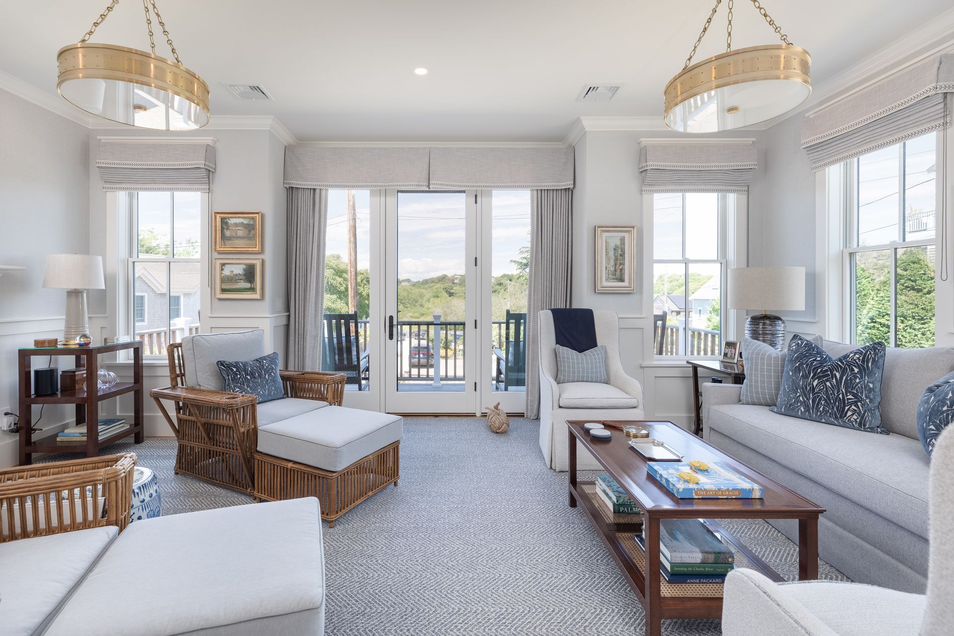 Bright coastal living room with blue-gray sofas, wicker chairs, and large windows overlooking water.