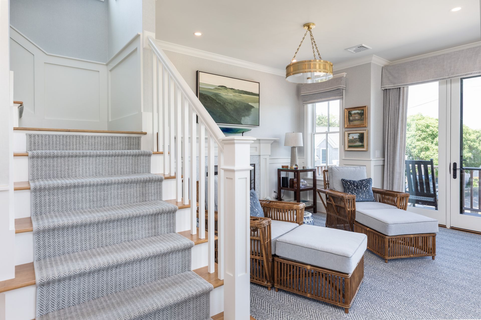 Bright living room with staircase, wicker seating, large windows, and coastal decor