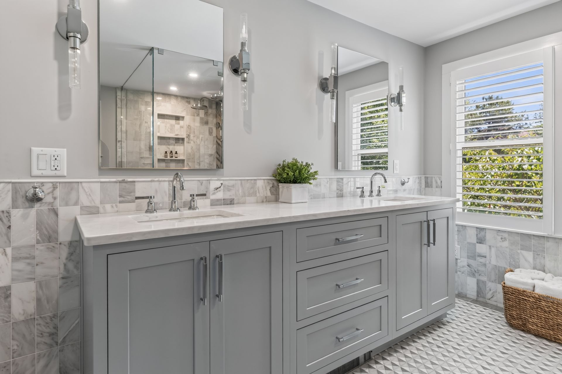 Bright gray bathroom with double vanity, large mirror, and patterned tile floor near a window and wicker basket