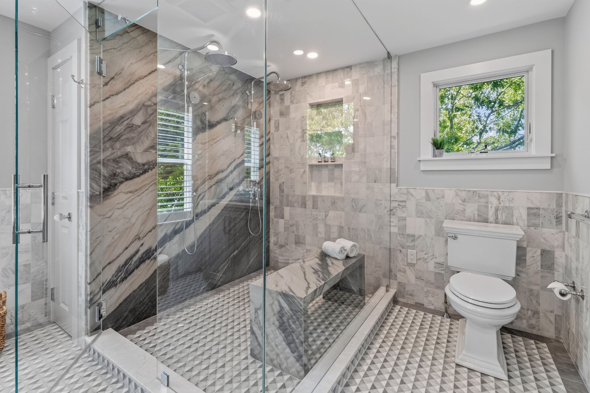 Bright modern bathroom with glass shower, marble walls, mosaic tile floor, and toilet by a window