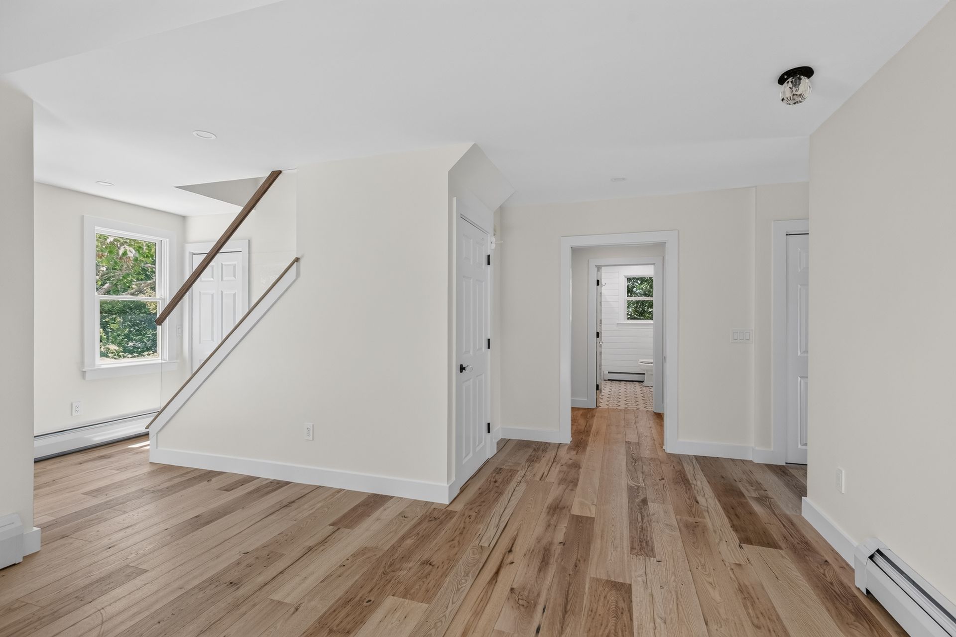 Bright empty hallway with wood floors, white walls, staircase, and doorway to another room