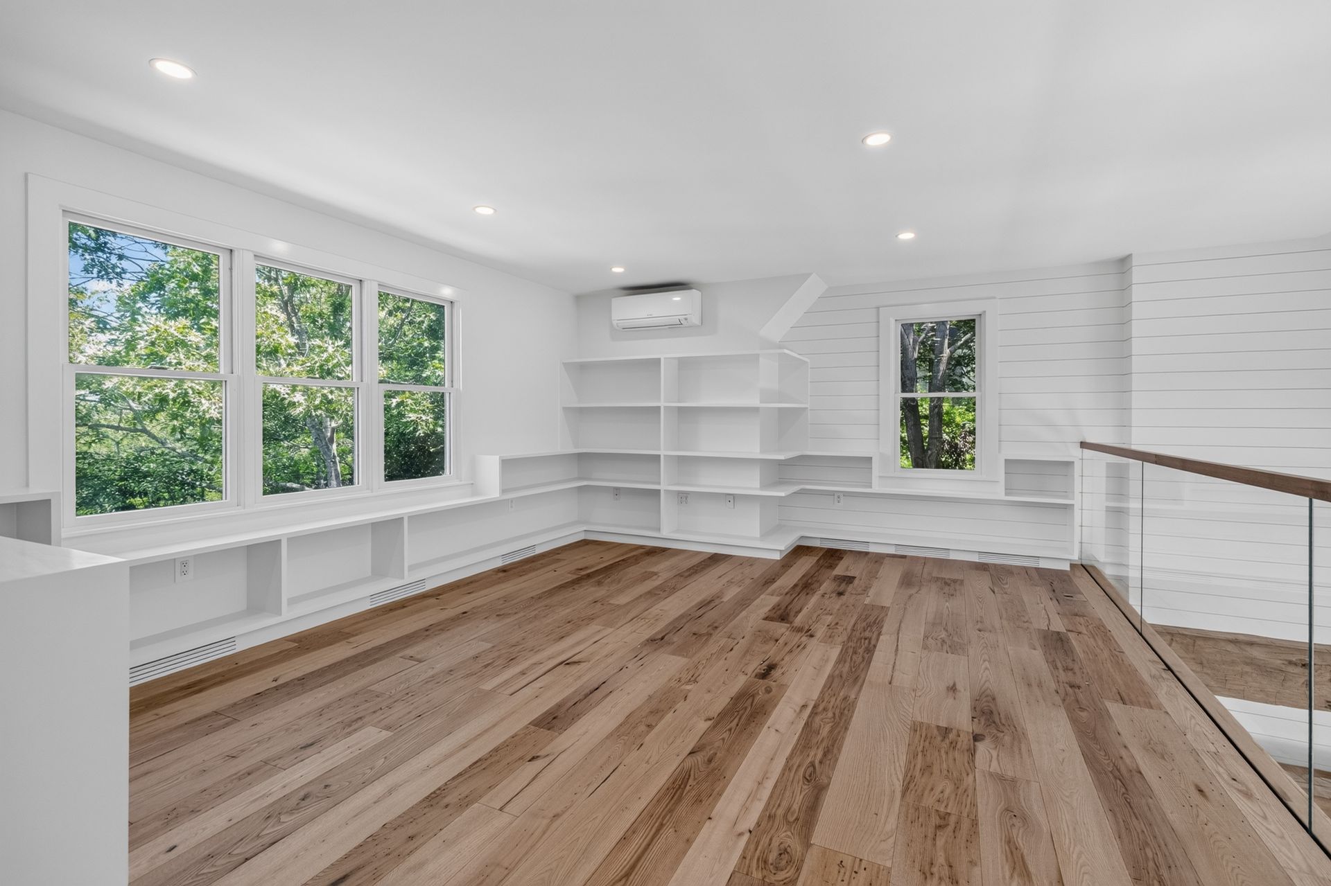 Bright empty room with wood floors, white walls, built-in shelves, windows, and recessed lighting