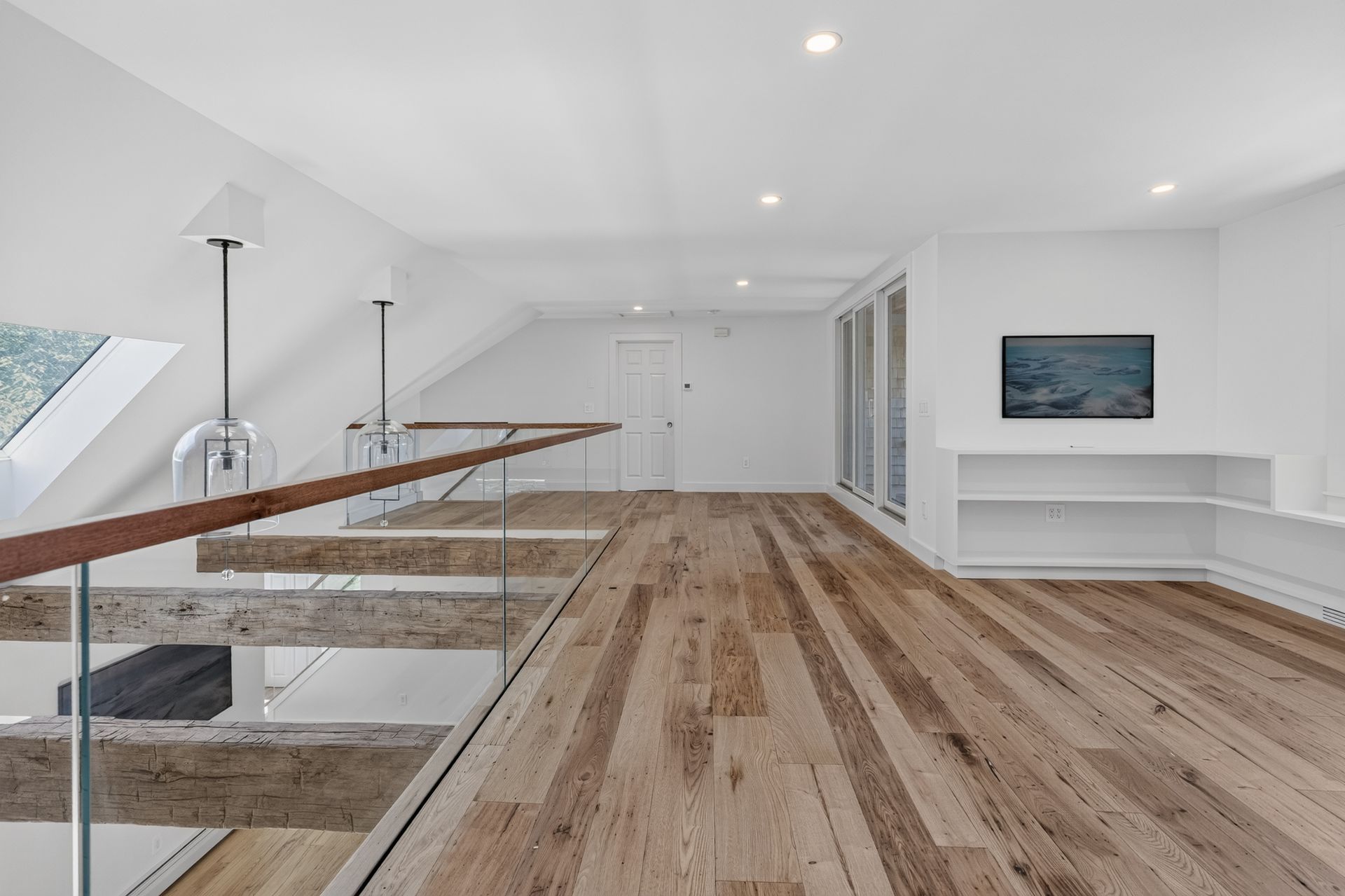 Bright modern hallway with hardwood floors, glass railing, and wall-mounted TV in a white interior room