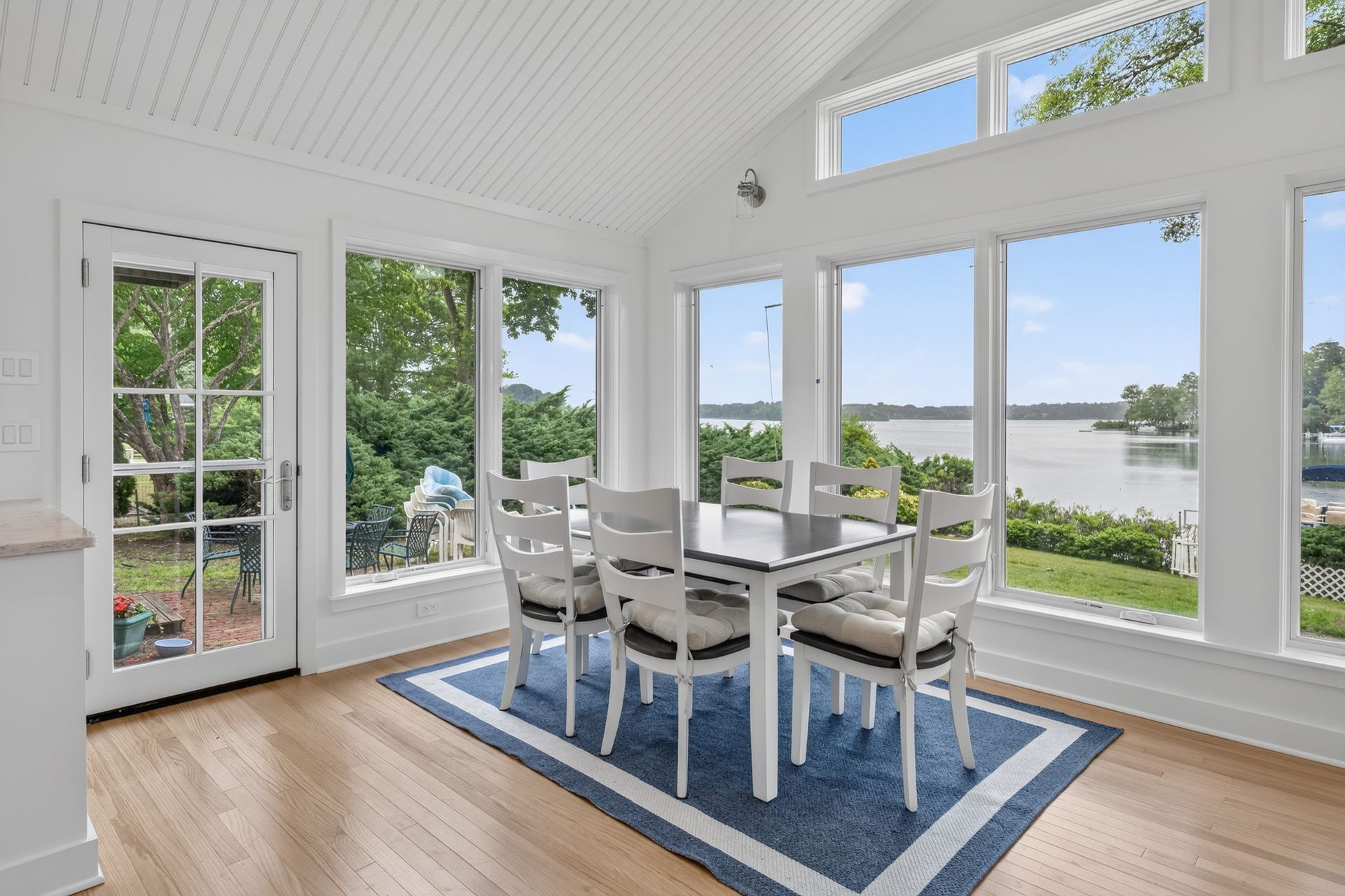 Bright dining room with white furniture, blue rug, and large windows overlooking the water