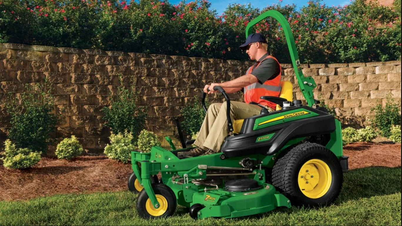 Man mowing a lawn on a green riding mower beside a stone wall and shrubs