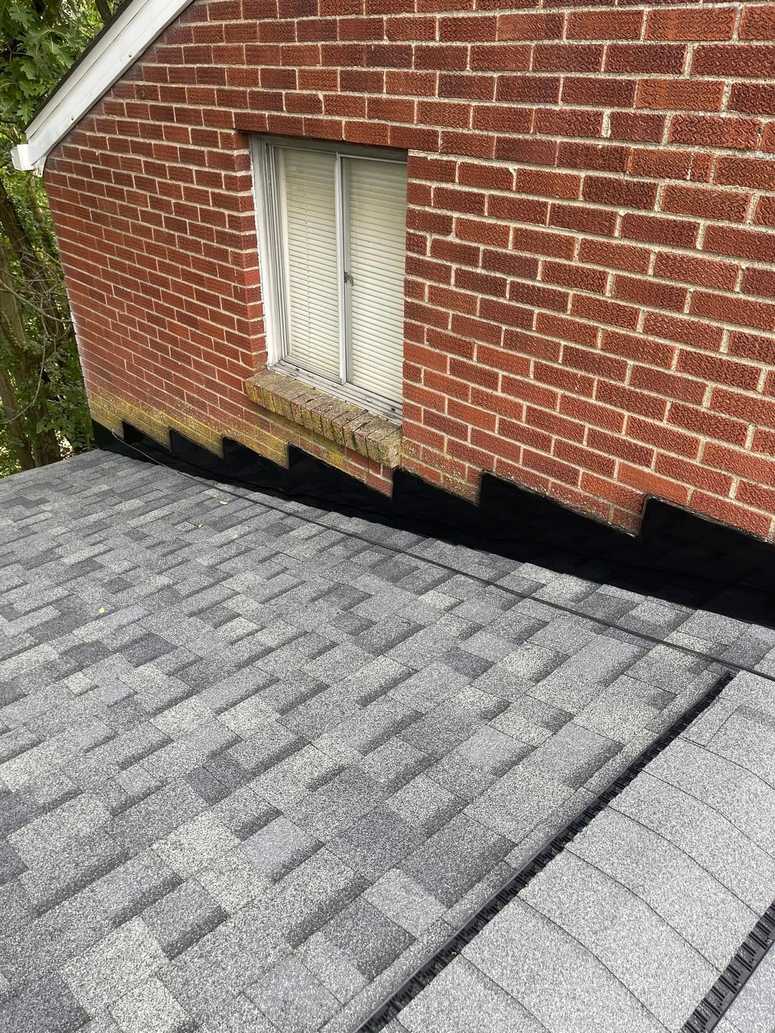 A gray asphalt shingle roof meets a red brick wall. A small window is visible in the brick wall.