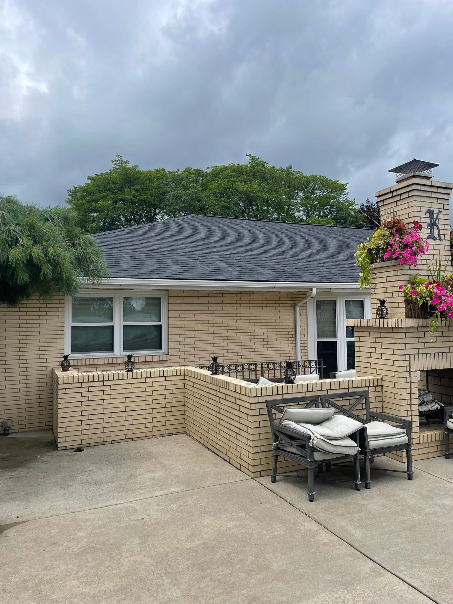 Backyard patio with a brick fireplace and seating area. Beige-colored house with black roof, overcast sky.