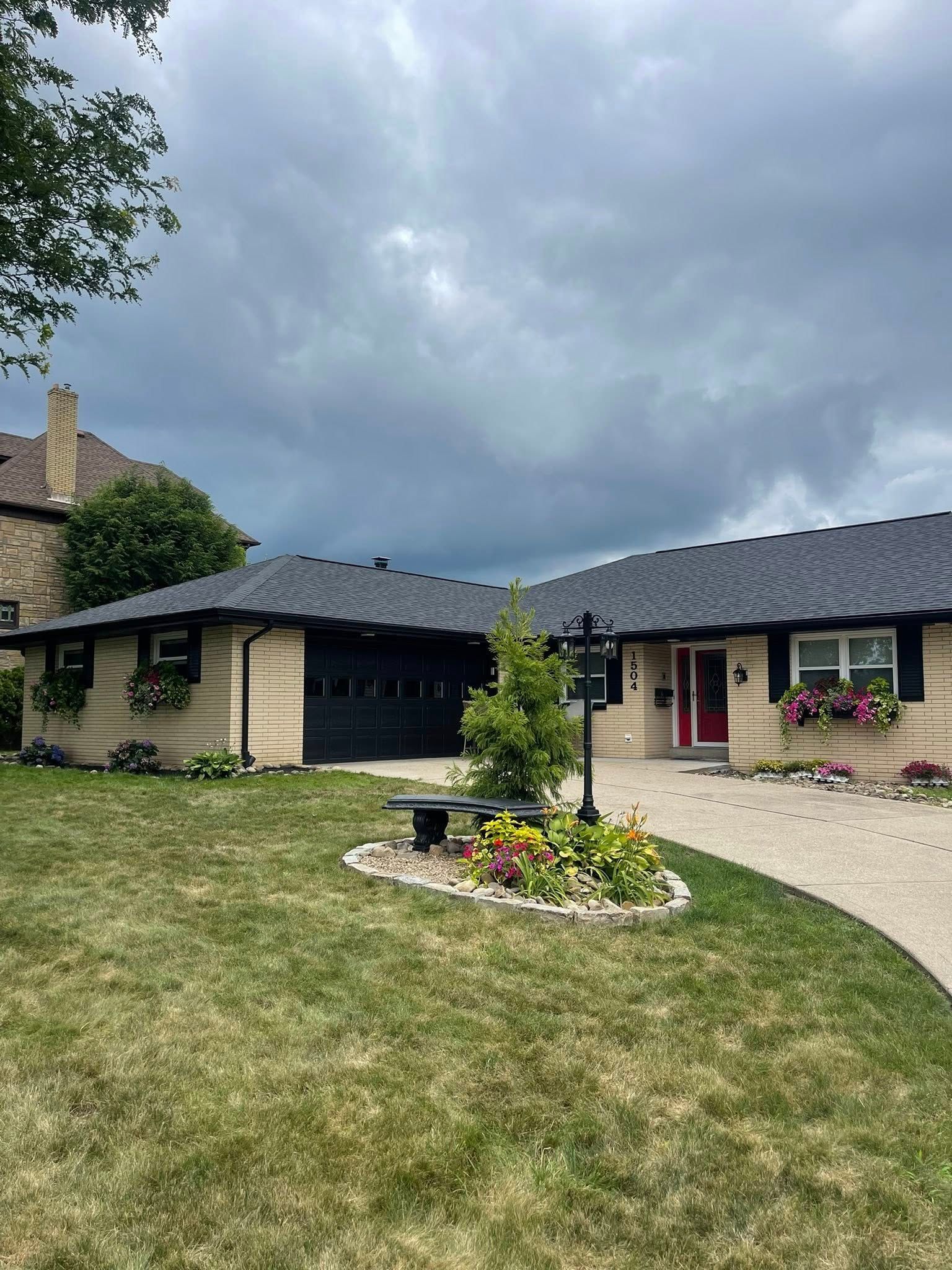 A tan ranch-style house with a black garage door and roof.  The front door is pink. There's a curved driveway and a dark, cloudy sky.