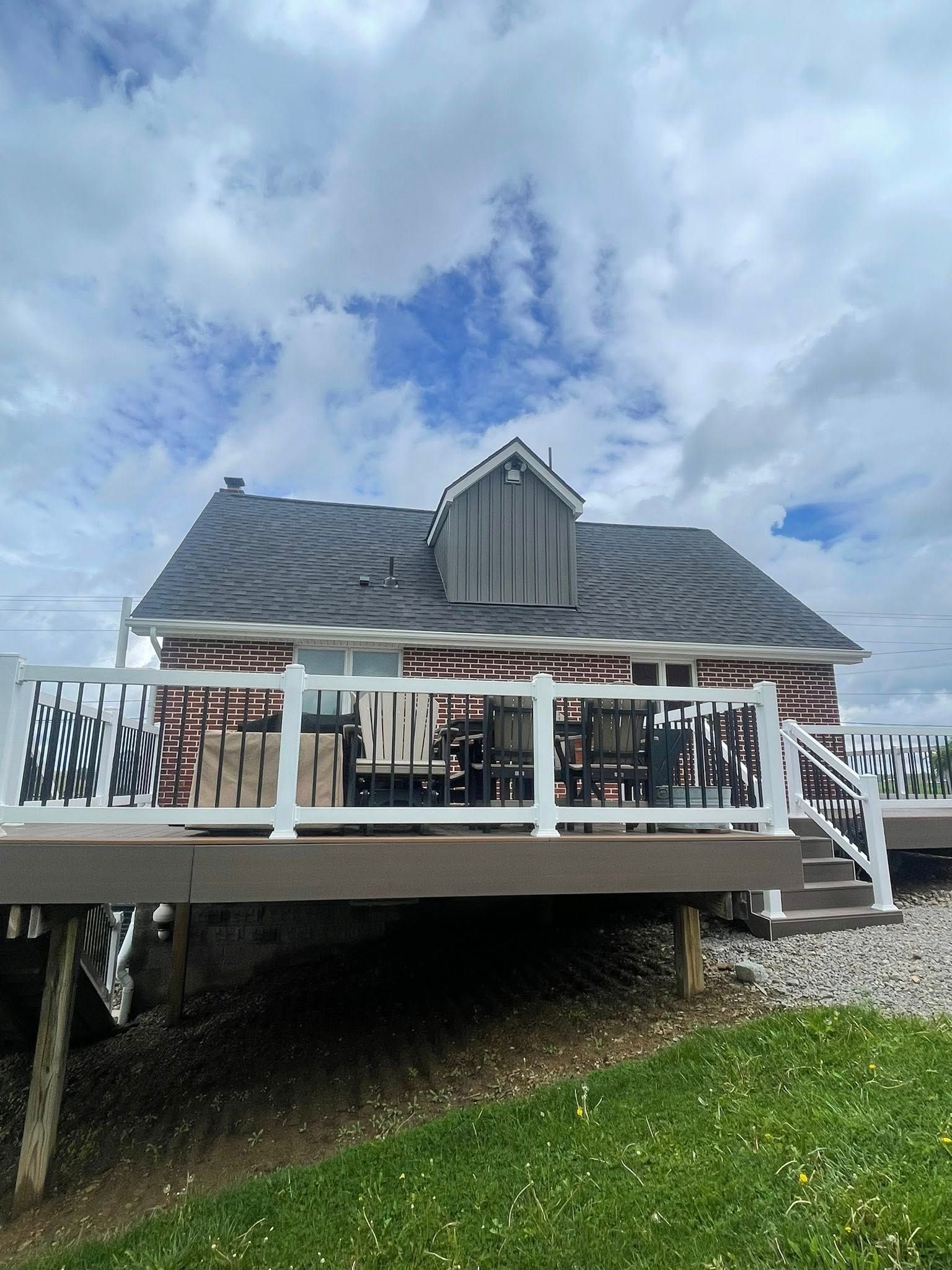 A house with a deck, gray roof, and red brick, under a cloudy sky. White railing surrounds the deck.